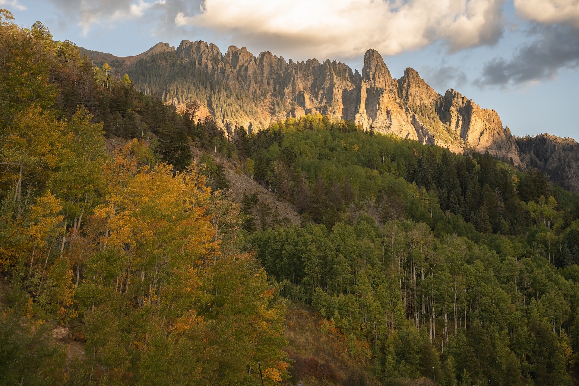 A mountain range with jagged peaks illuminated by sunlight, surrounded by a forest with green and autumn-colored trees, with a partly cloudy sky above.