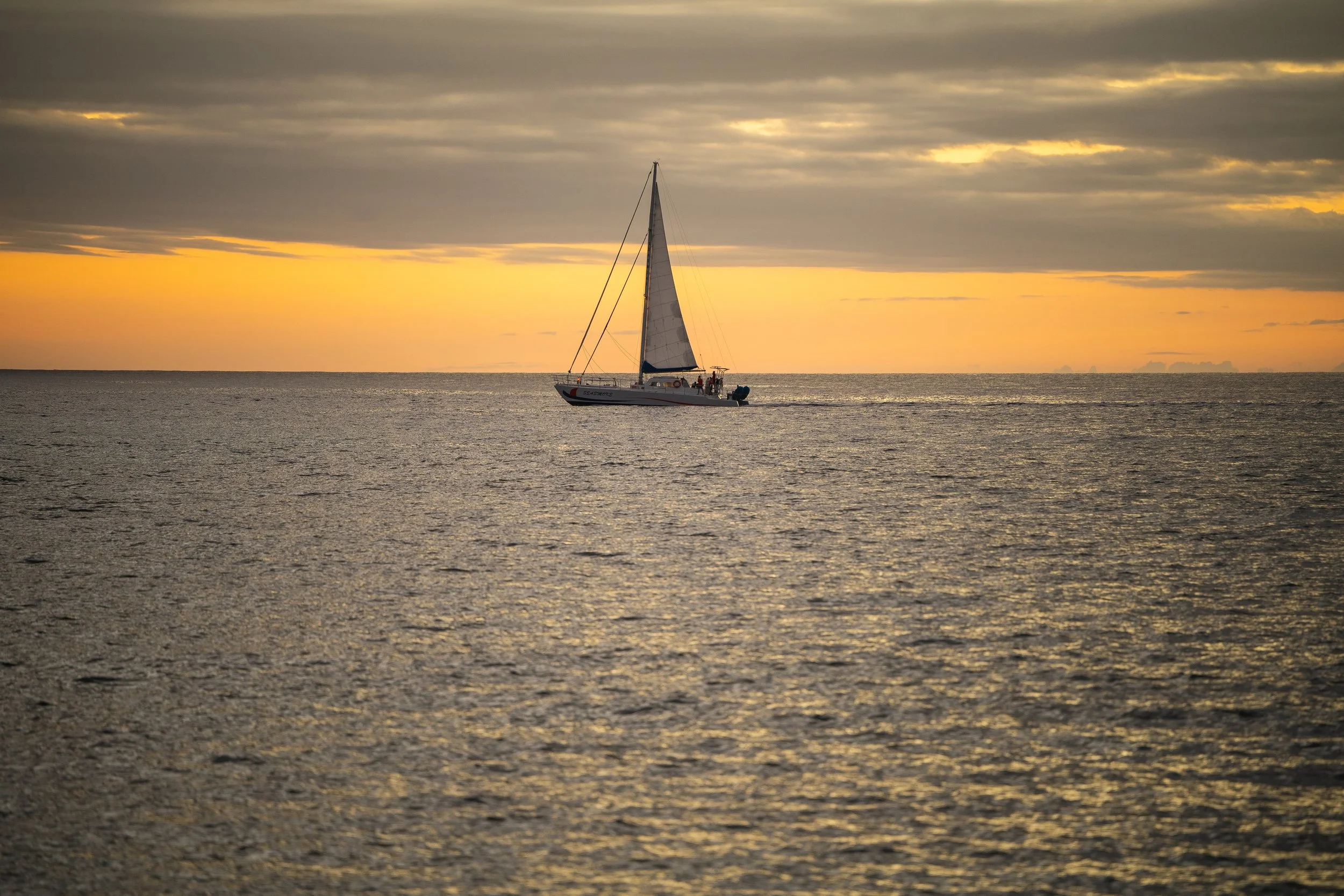 A sailboat sailing on calm ocean waters during a sunset with a partly cloudy sky.