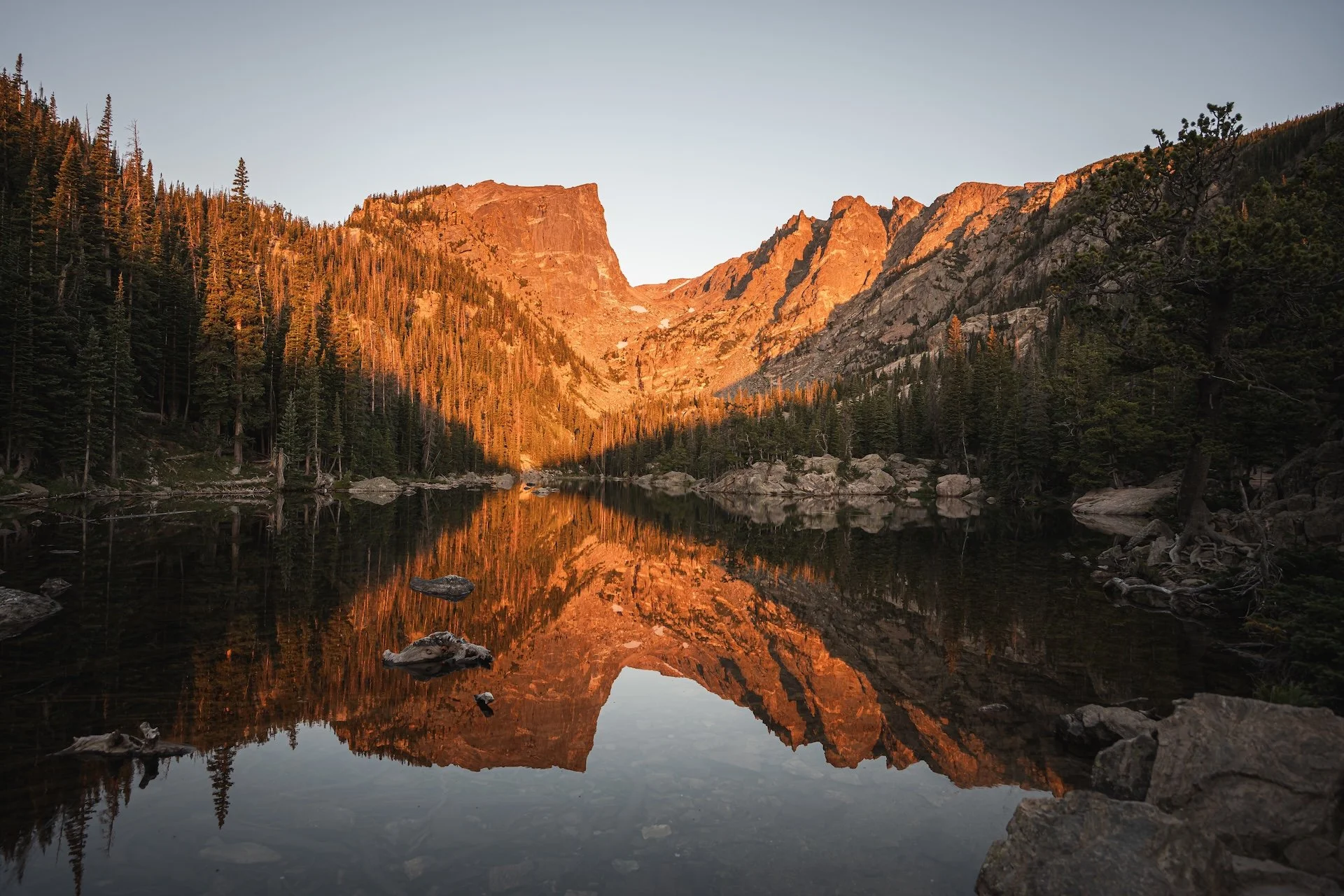 Mountains reflected in a calm lake at sunset, surrounded by pine trees and rocky shore.