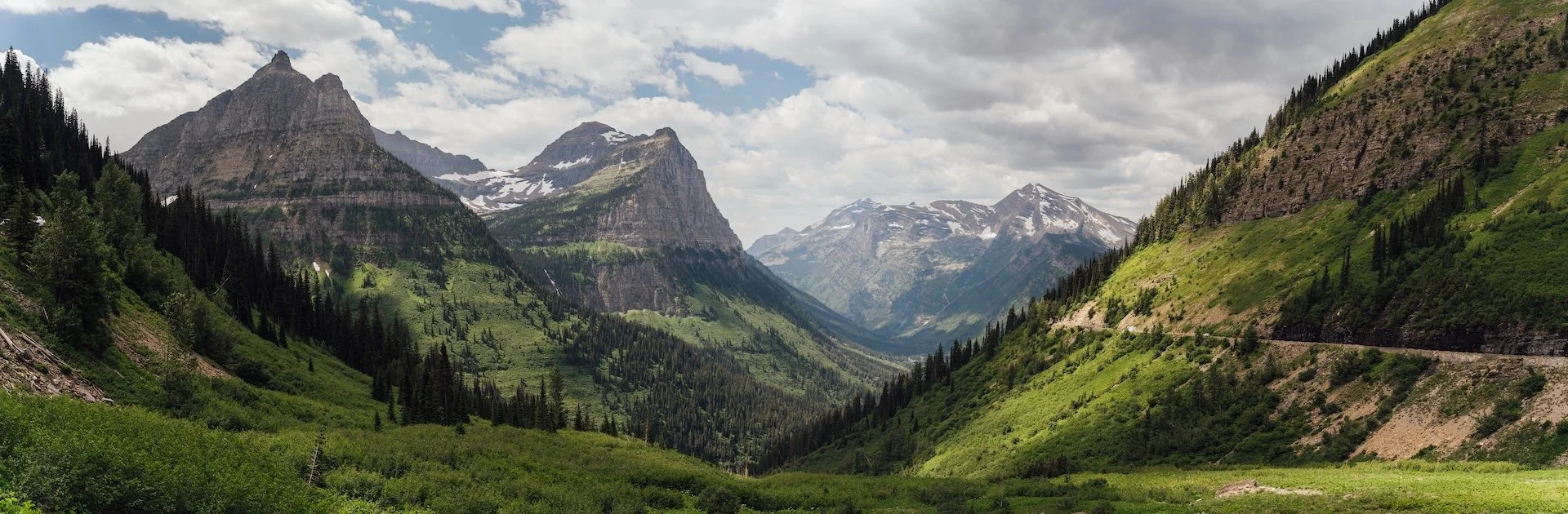 A lush green mountain valley with tall pine trees, rugged mountains with snow patches in the distance, partly cloudy sky.