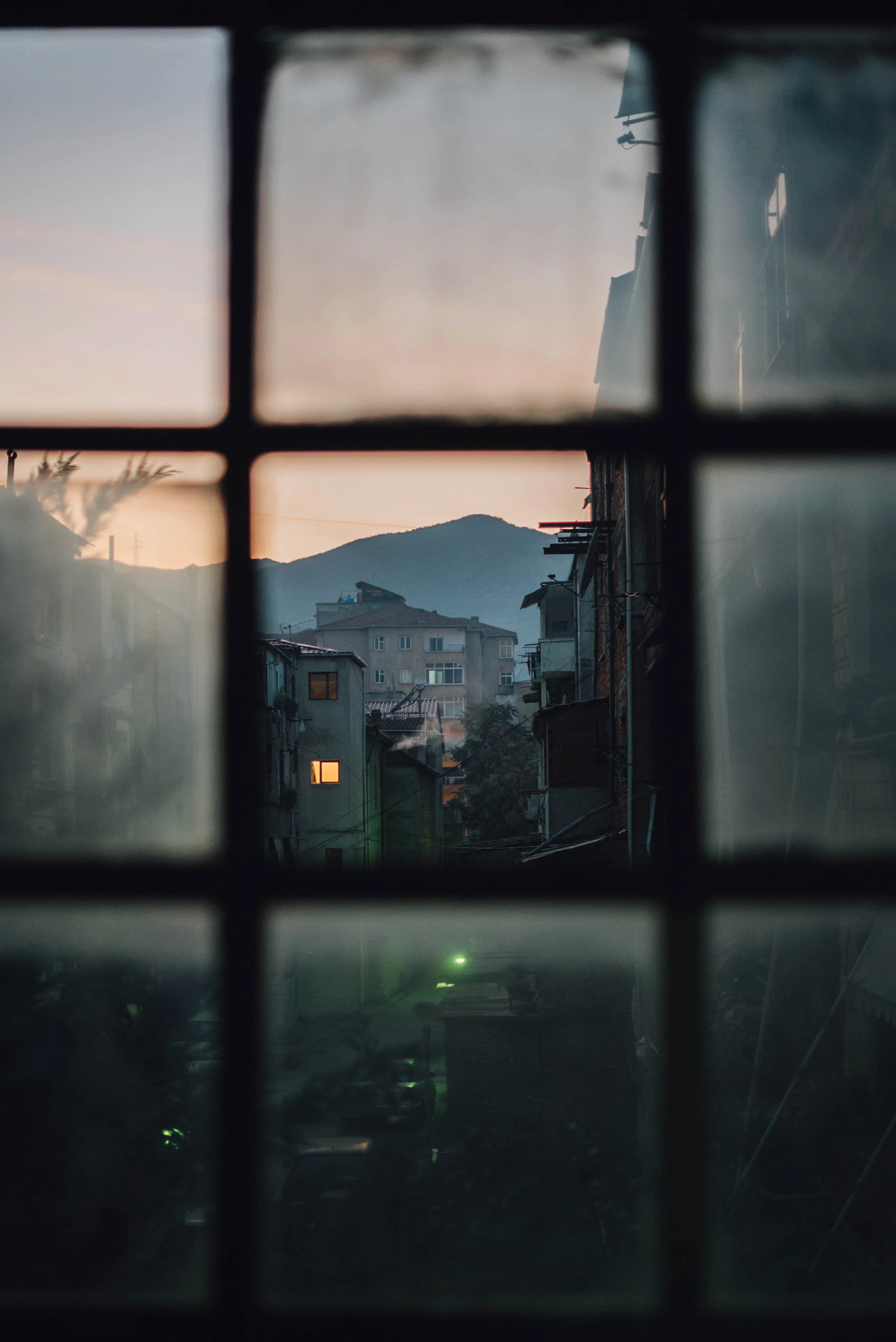View through a window, showing buildings, a lit window, and a mountain at sunset.