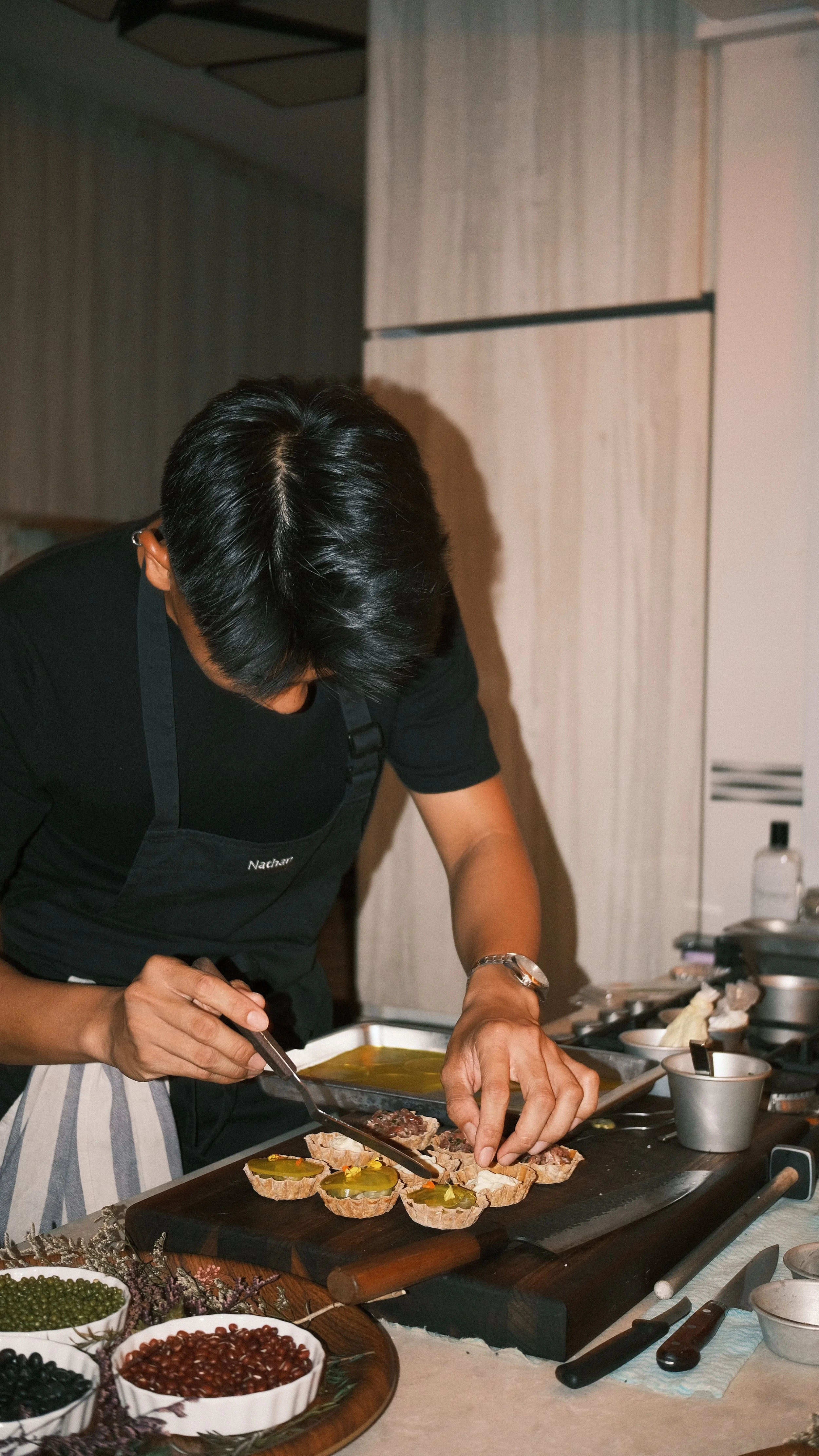A person preparing food, slicing a small piece of meat or bread, on a slate board with toppings. Various bowls with ingredients like peppercorns are on the table, with kitchen utensils and containers in the background.