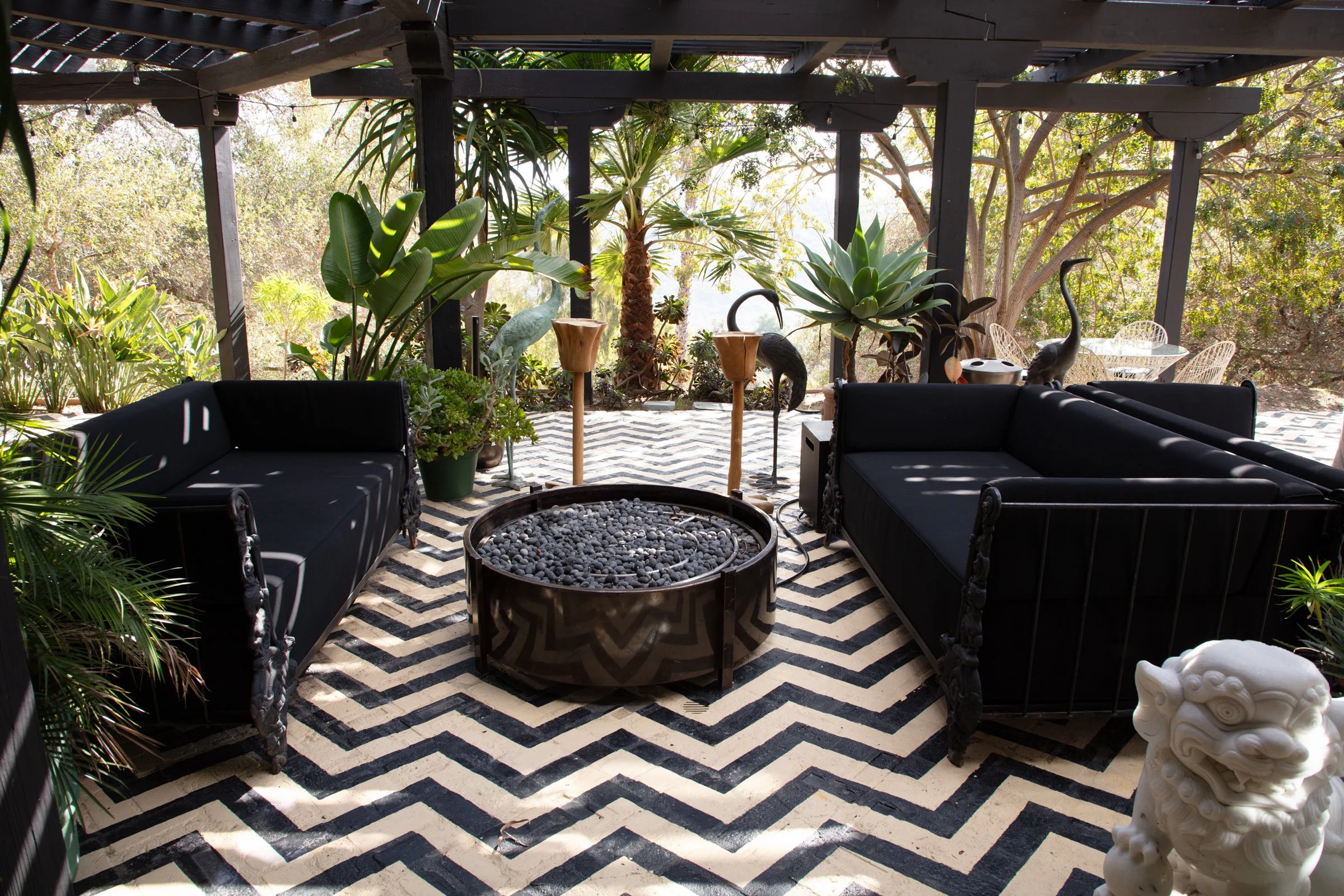 Outdoor patio with black sofas, large potted plants, a patterned black and white rug, decorative flamingos, a fire pit filled with black stones, and patio furniture in the background.