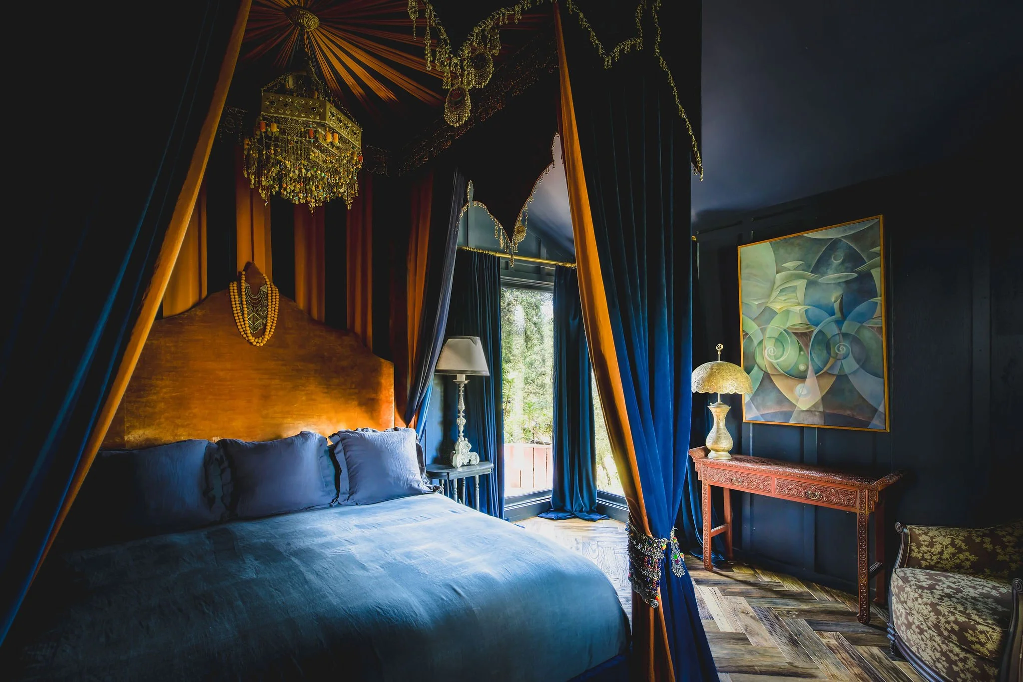 Luxurious bedroom with dark blue walls, a four-poster bed with blue bedding, gold and blue curtains, vintage lamps, and artwork on the wall, with natural light coming through a nearby window.