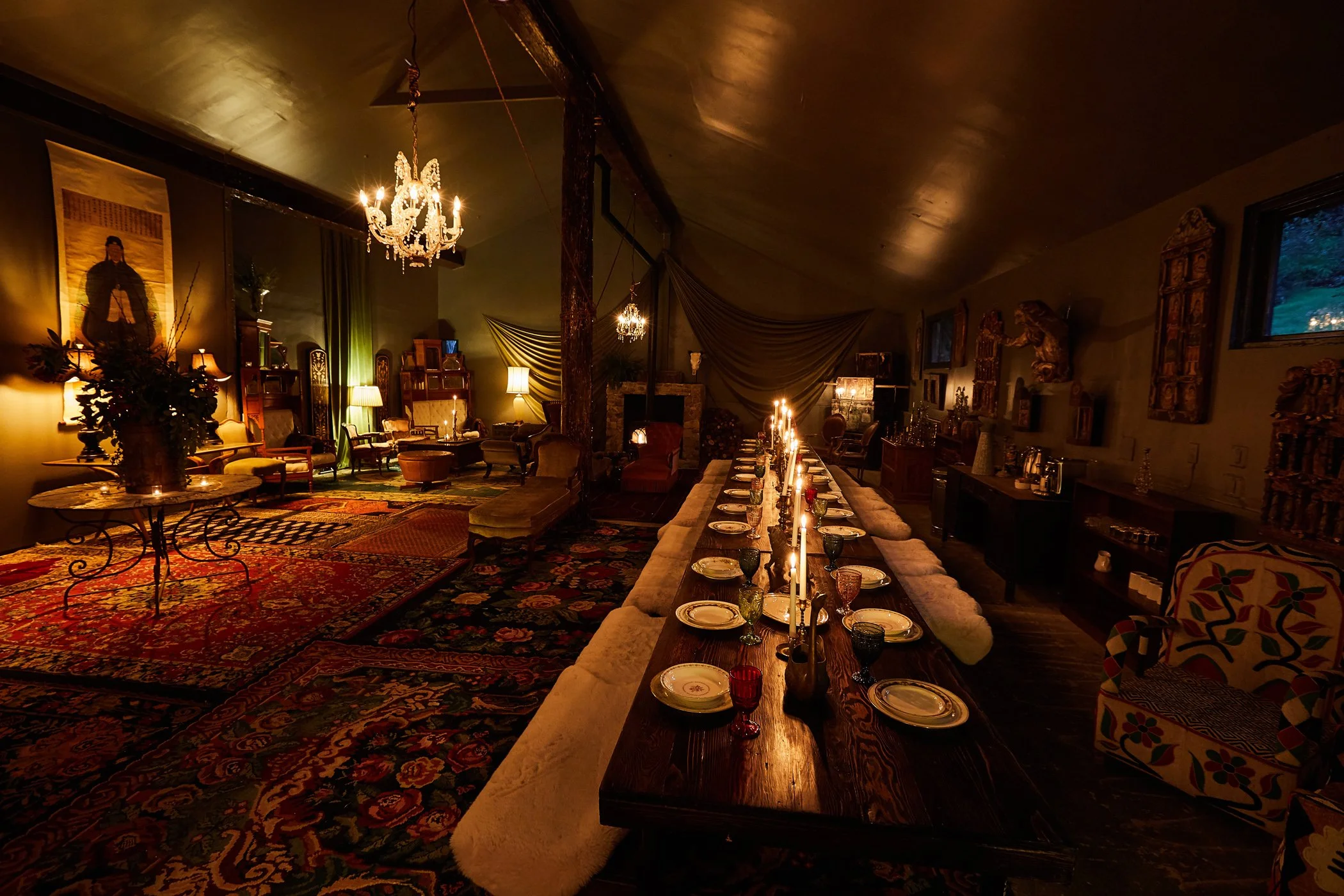 Dimly lit vintage dining room with a long wooden table set with plates, glasses, and candles, surrounded by cushioned chairs, ornate rugs, chandeliers, antique furniture, and decorative artwork on the walls.