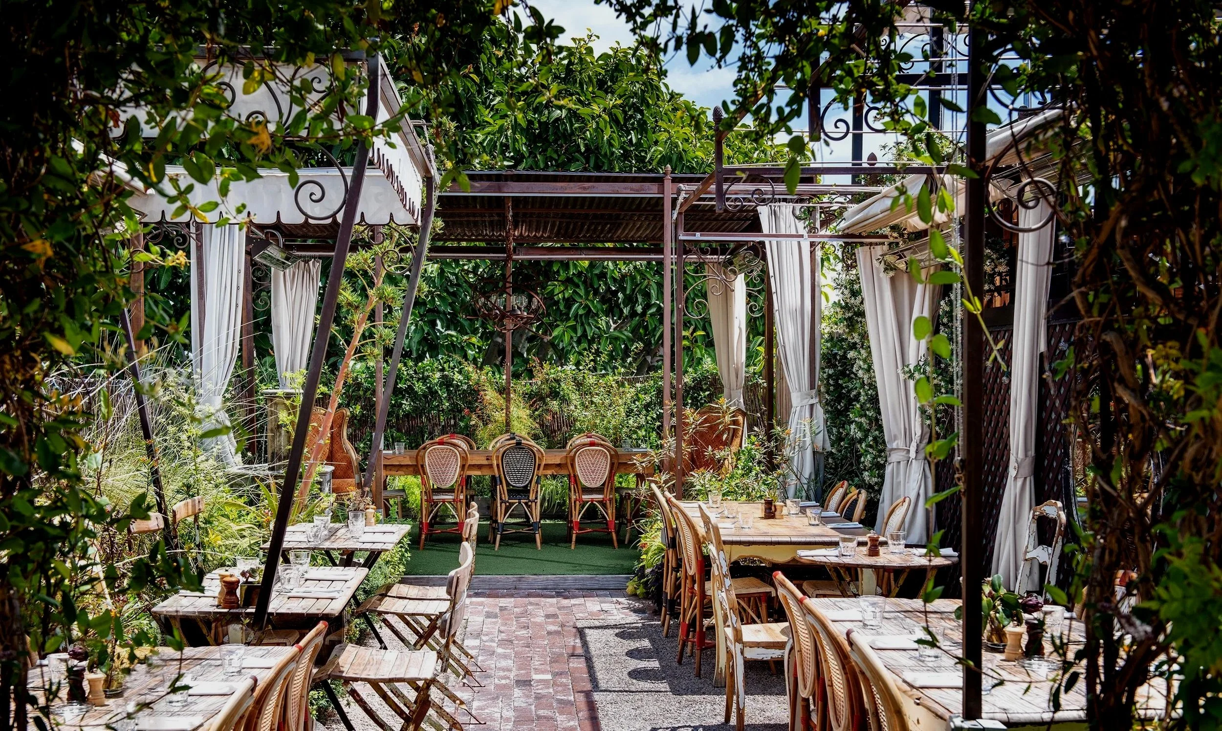 Outdoor restaurant patio with wooden tables and chairs, surrounded by lush green plants and trees with white curtains hanging from a pergola.