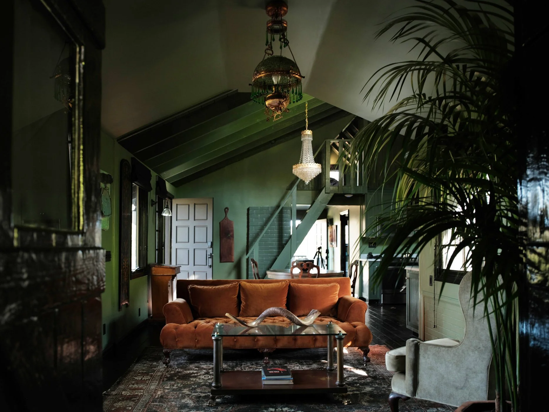 Cozy living room with green walls, vintage decor, an orange velvet sofa, a glass coffee table with decorative items, a white armchair, hanging lights, a staircase, and a large potted plant.