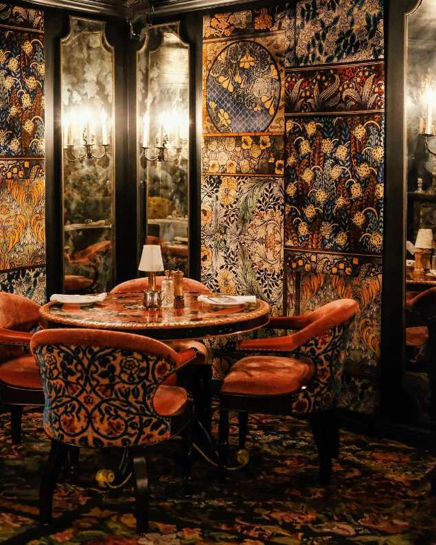 Elegant dining room with ornate wallpaper, a round table set for four, velvet upholstered chairs, mirrored walls, and soft lighting.