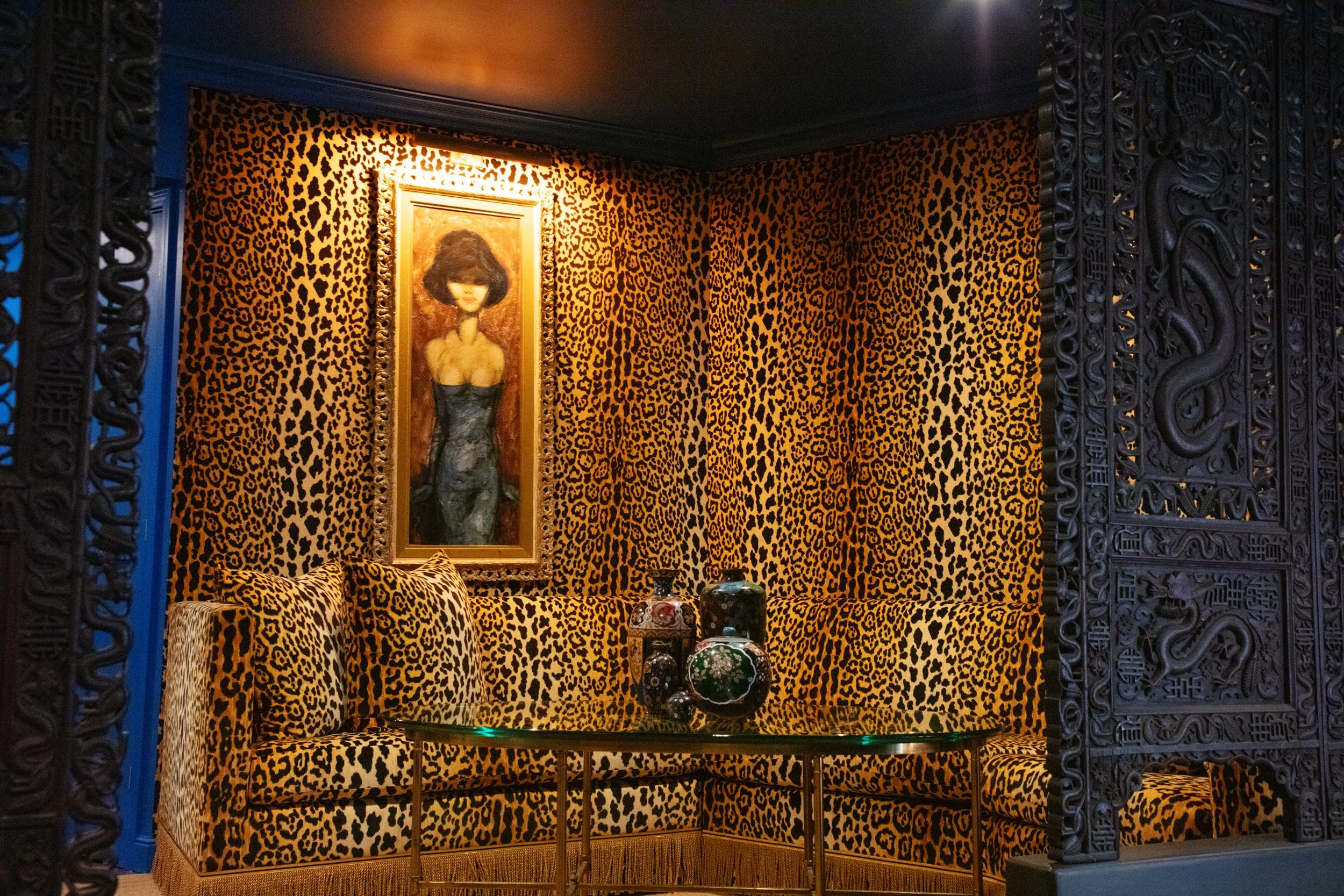 A room with animal print (leopard) patterned walls and matching furniture, a painting of a woman with dark hair, and a glass table with decorative vases. Ornate black carved wood framing surrounds the scene.