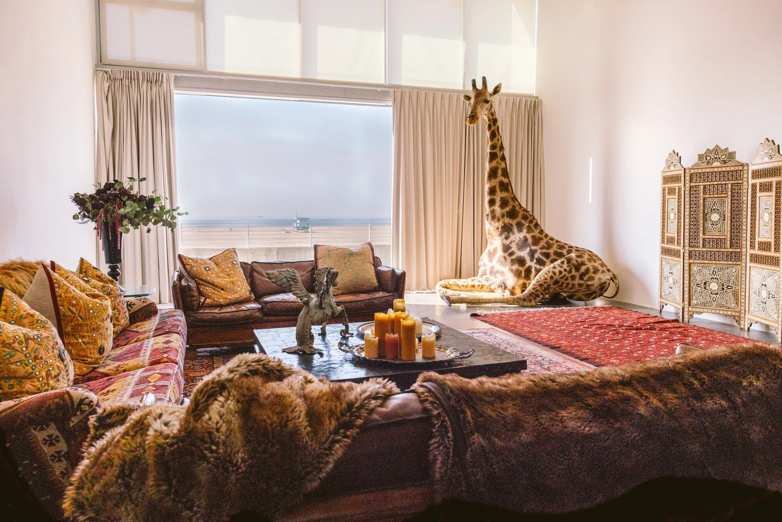 Living room with eclectic decor, featuring a large giraffe sculpture near a window, a small winged horse statue on a coffee table, and couches with patterned cushions. There is a decorative screen on the right and an ocean view through the window.