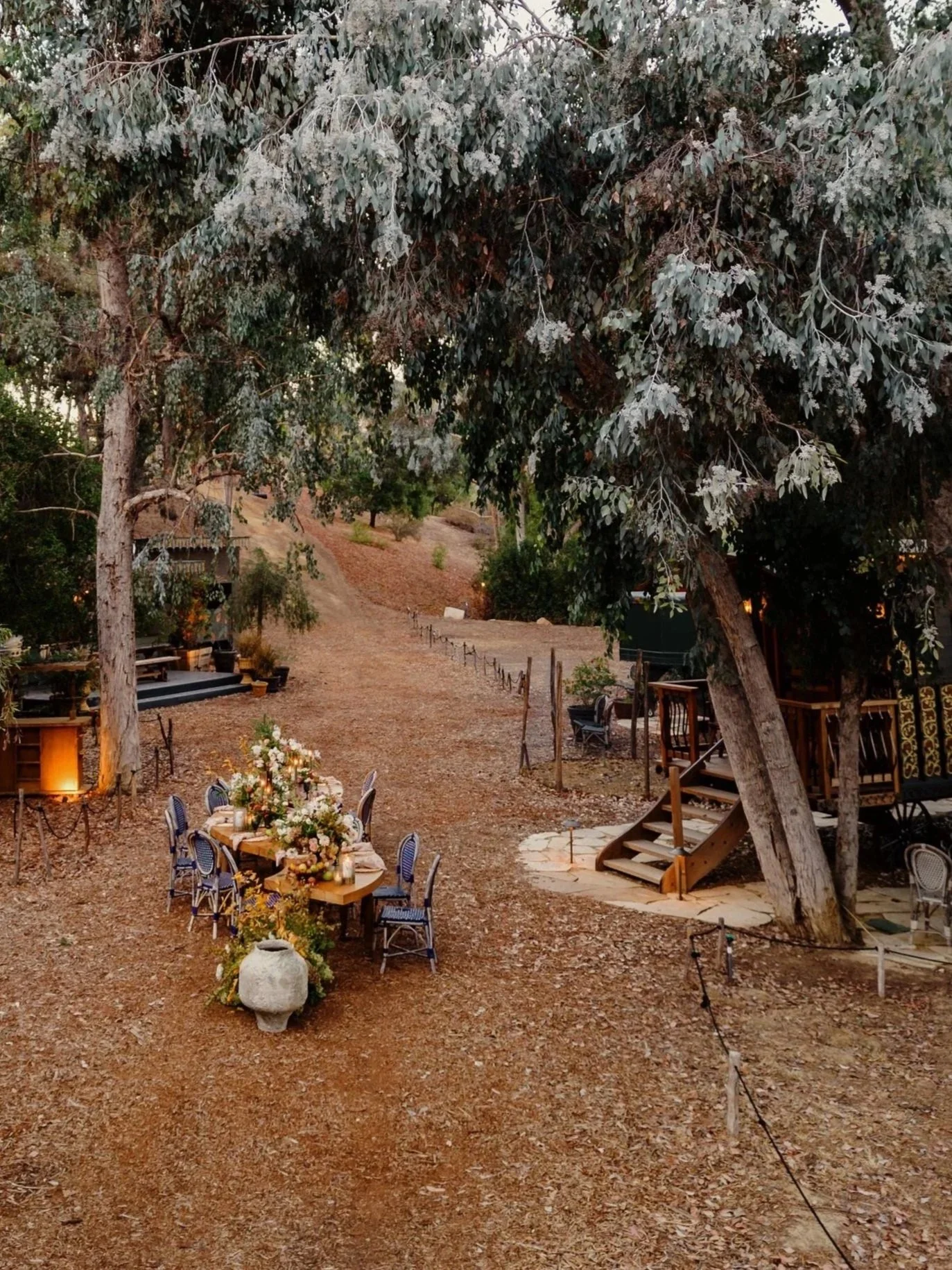 An outdoor setting with a long table decorated with flowers and surrounded by chairs, situated on a dirt path under large trees, in what appears to be a rustic or garden venue ready for an event.