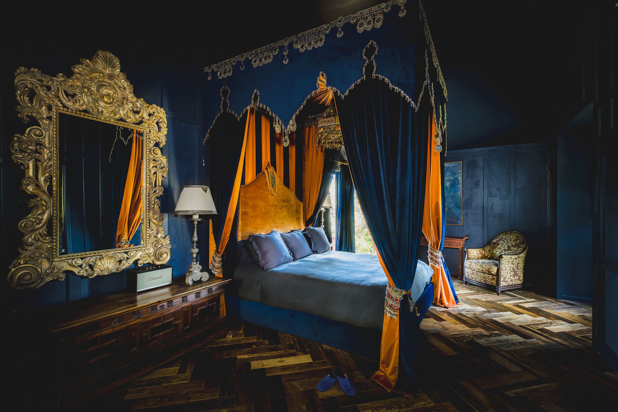 Luxurious bedroom with dark blue walls, ornate gold-framed mirror, four-poster bed with blue and orange curtains, a bedside lamp, an upholstered armchair, and a window with natural light.