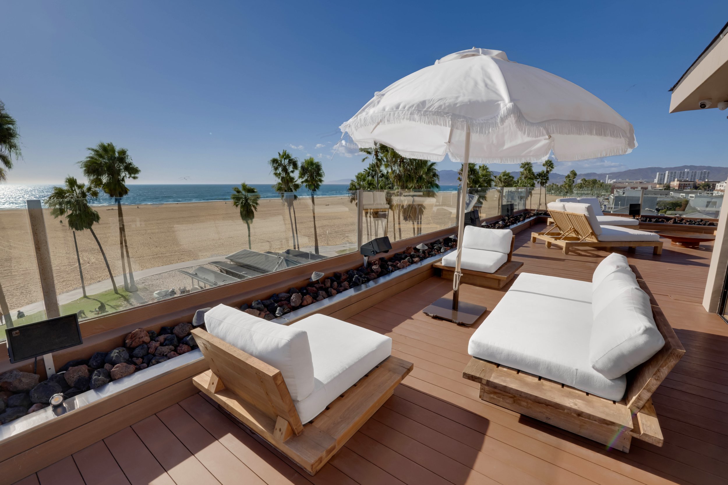 Beachside outdoor lounge with white cushioned wooden furniture, a large white umbrella, and a view of palm trees and the ocean in the background.