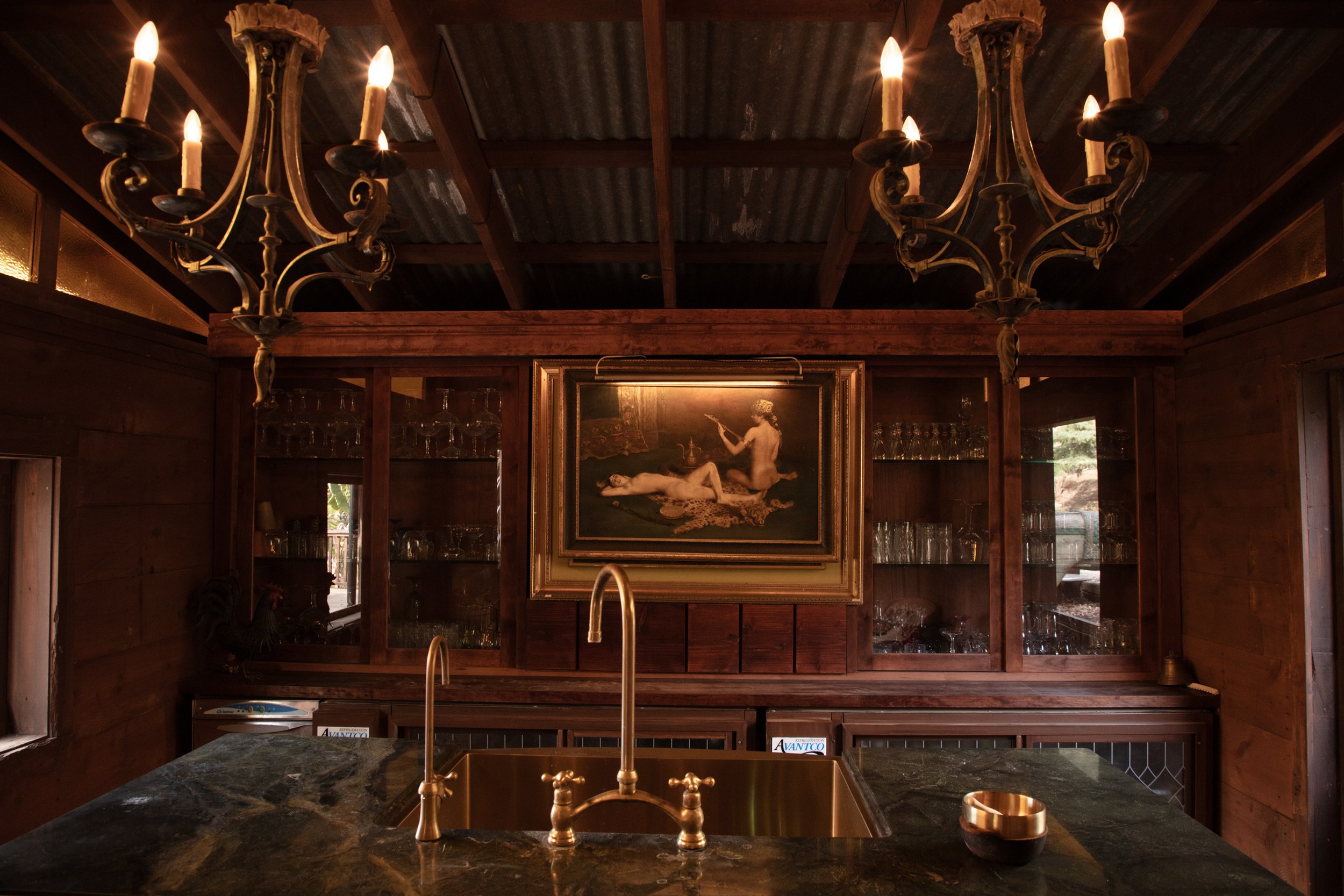 A rustic wooden kitchen or bar area with two elegant chandeliers, a painting of two nude women, a marble countertop with a gold-colored sink, and glassware displayed in cabinets.