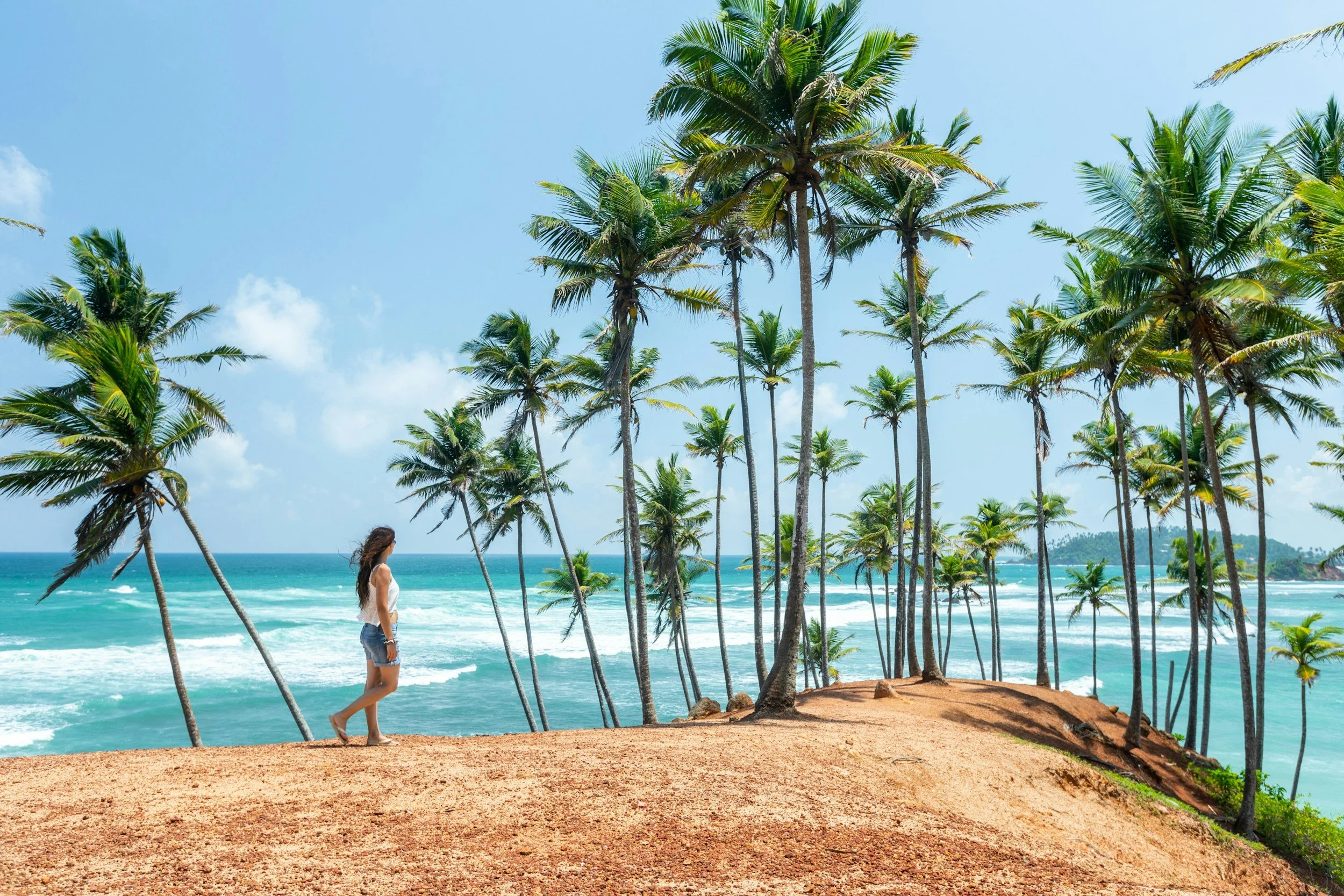 A woman standing on a sandy hill on a tropical beach with tall palm trees, turquoise water, and a blue sky with a few clouds.