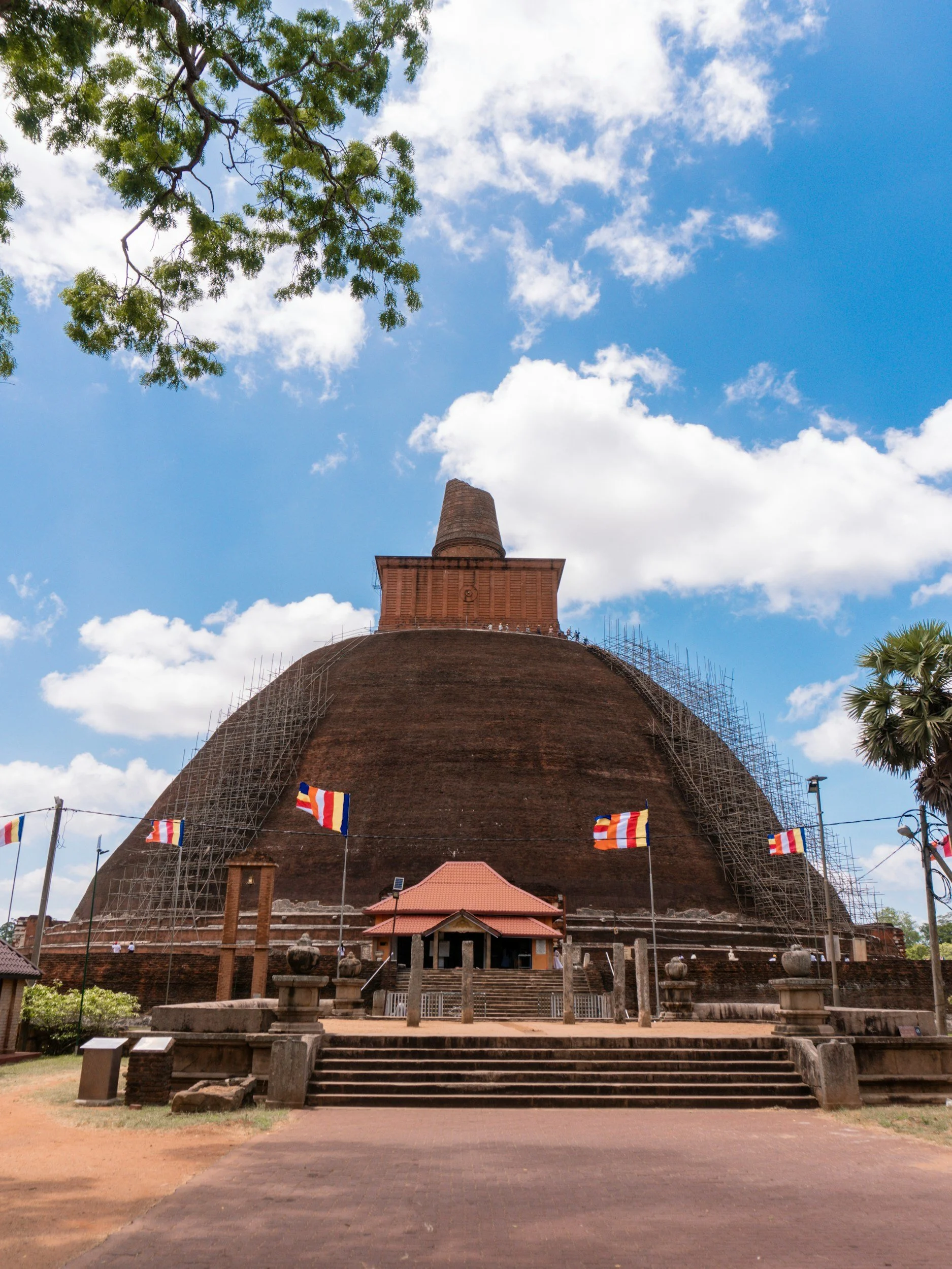 A large, ancient stupa with scaffolding on its sides, under a bright blue sky with scattered clouds, surrounded by a few trees and flags.