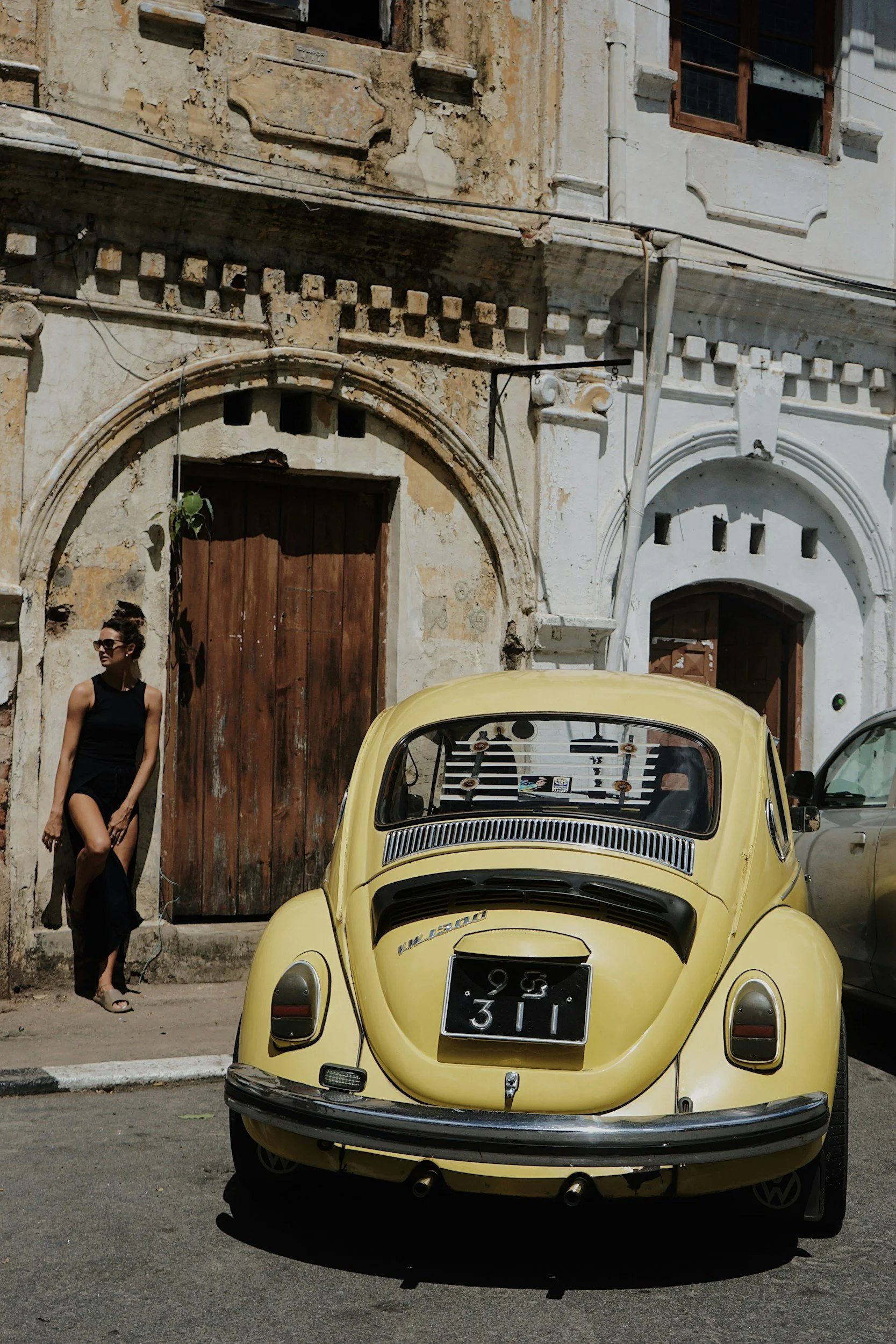 A woman in a black dress standing next to a yellow vintage Volkswagen Beetle with an old building in the background.