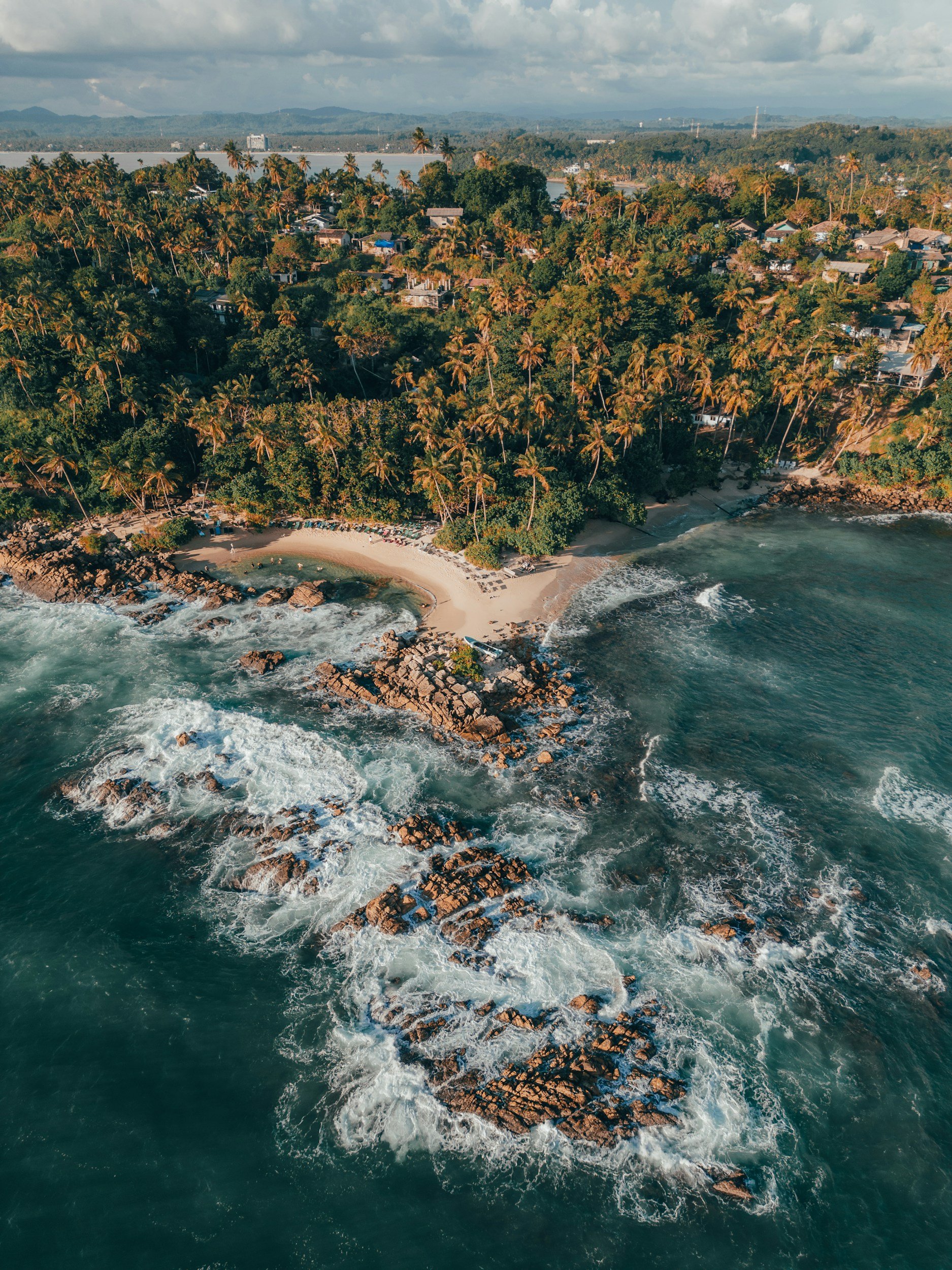 Aerial view of a tropical beach with rocks, waves, sandy shore, lush green trees, and palm trees, with a forest and distant mountains in the background.
