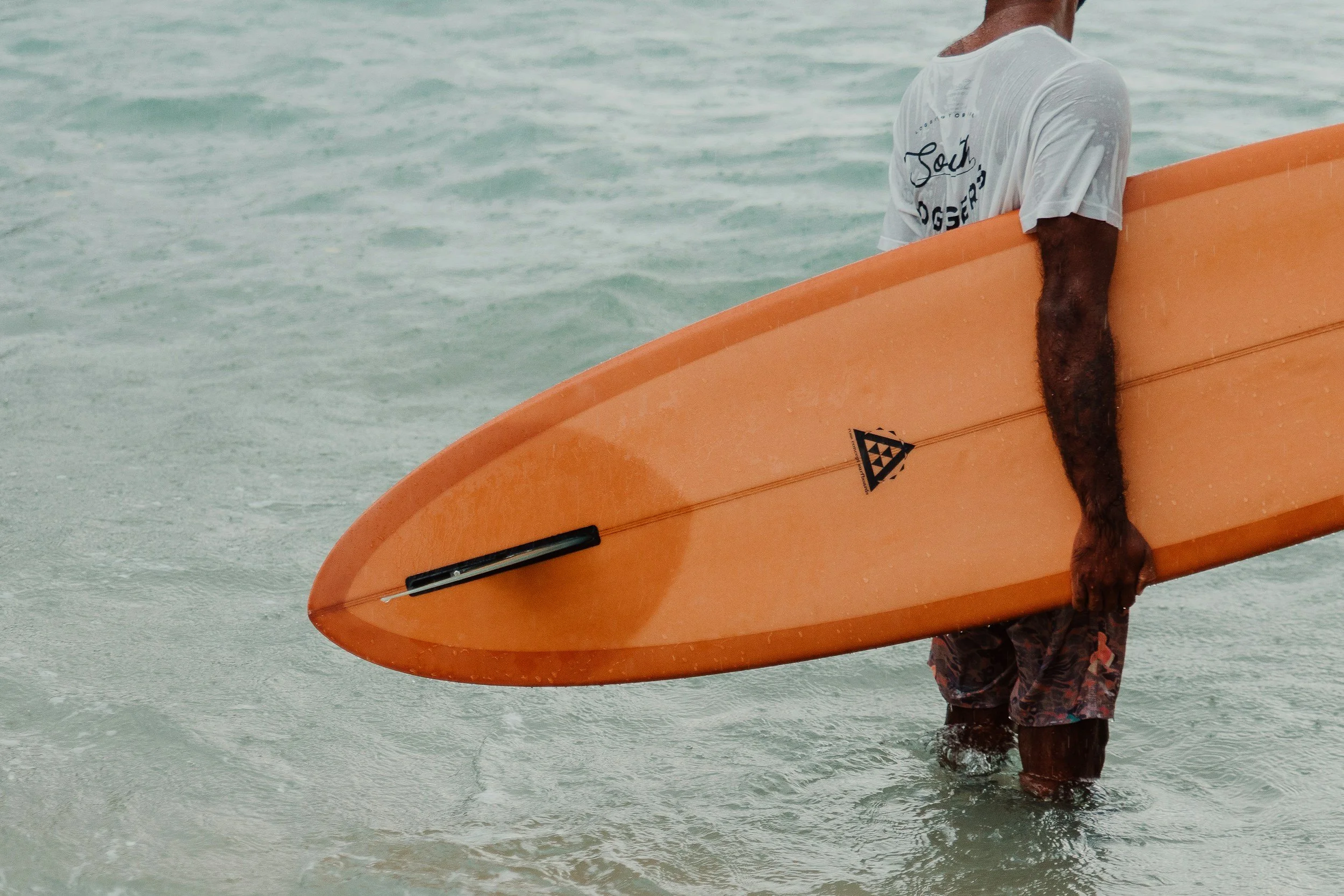 A person standing waist-deep in the water holding an orange surfboard with a black fin and leash, wearing a white T-shirt and patterned shorts.
