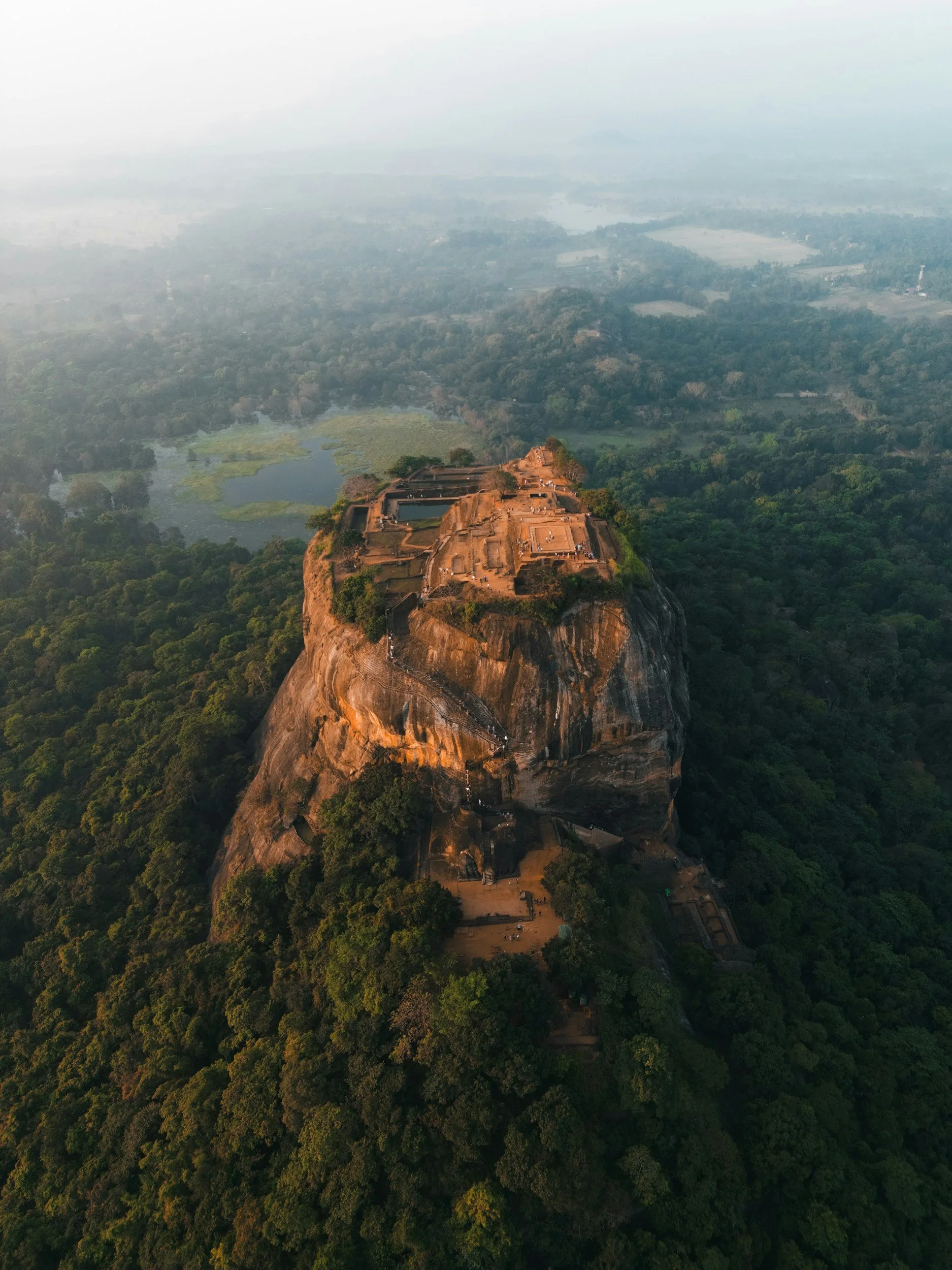 Aerial view of Sigiriya rock fortress surrounded by lush green forest and distant lakes.