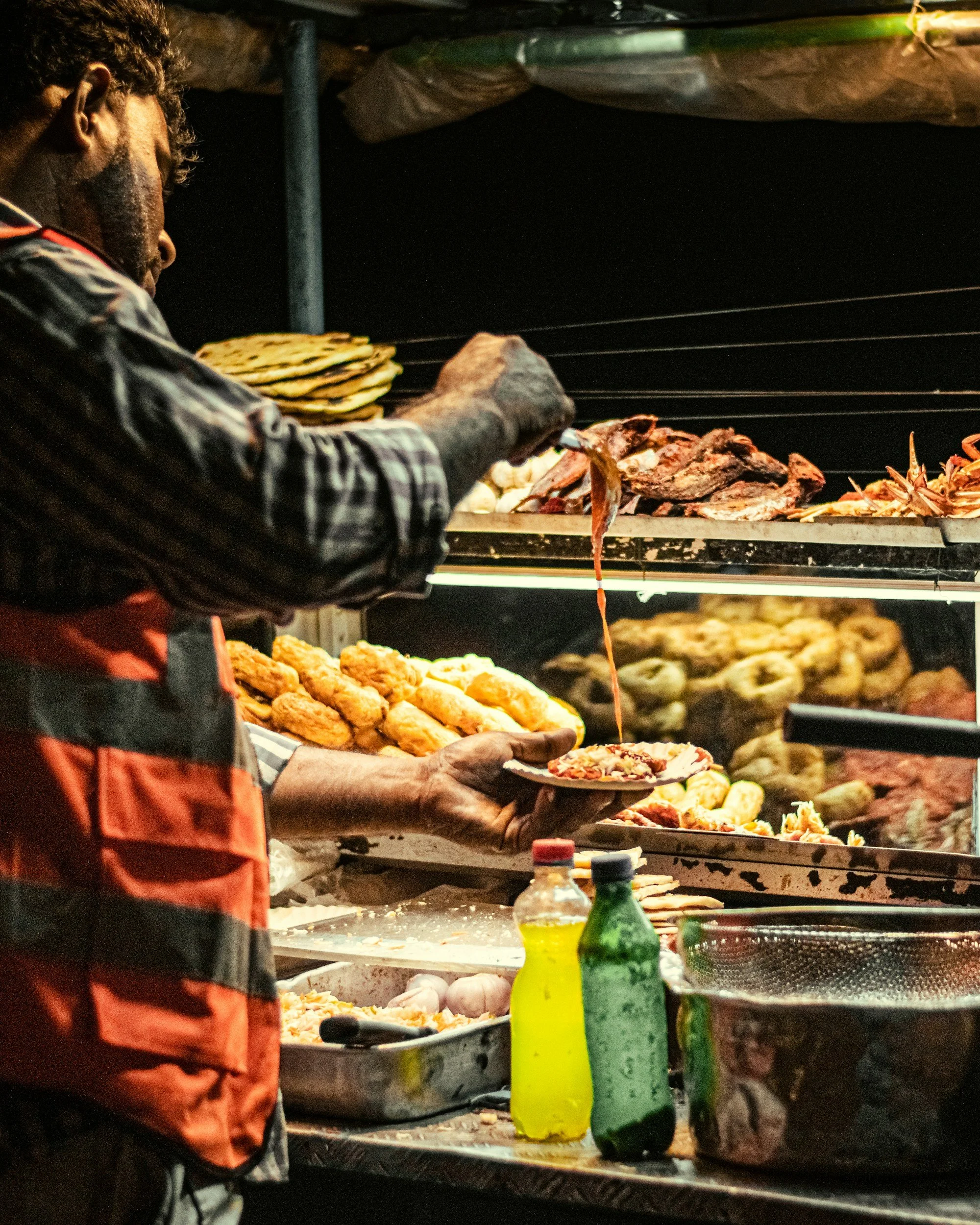 A man preparing and serving food at a street food stand with various grilled meats, fried foods, and condiments.