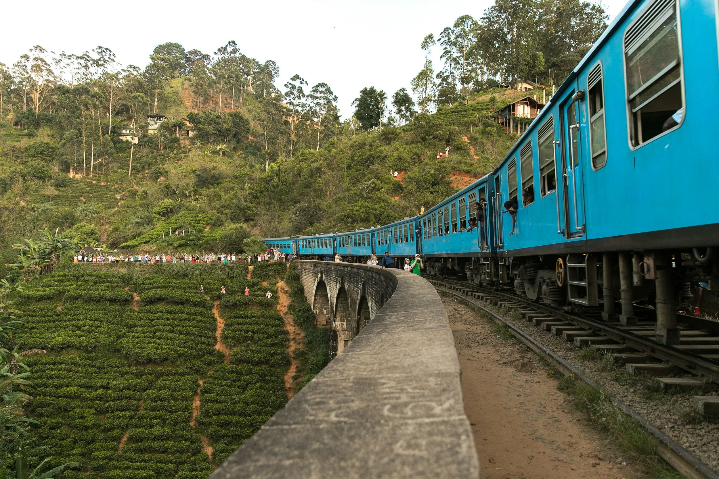 Blue train traveling around a mountain curve with passengers looking out from the windows, set against lush green hills and terraced fields.