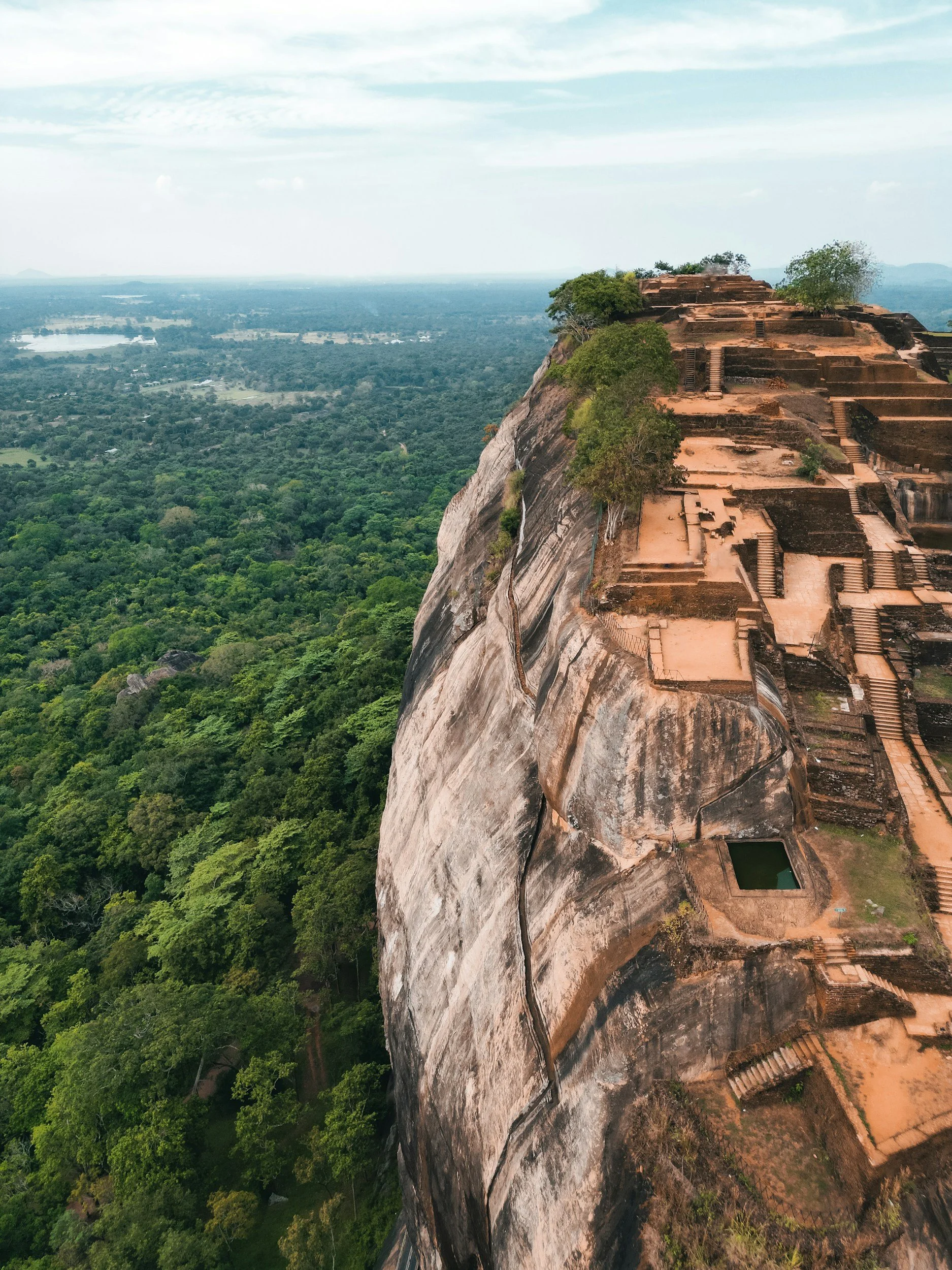 Ancient stone structures built on a steep rocky hill overlooking a green forest and a distant water body.