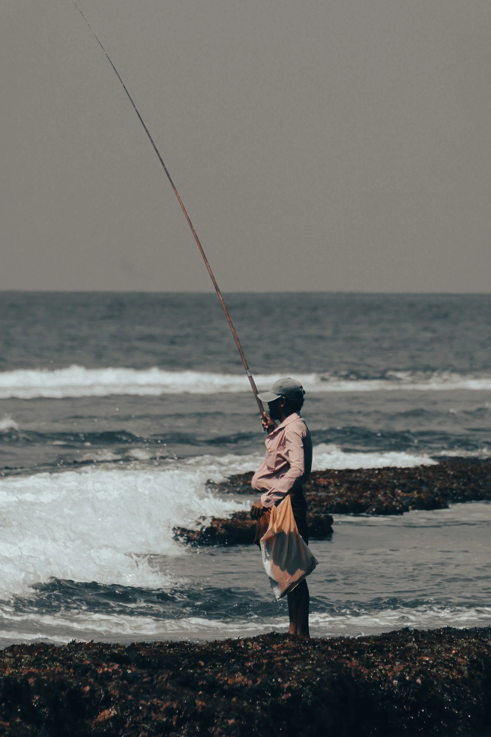 A person standing on rocks by the ocean, fishing with a fishing rod.
