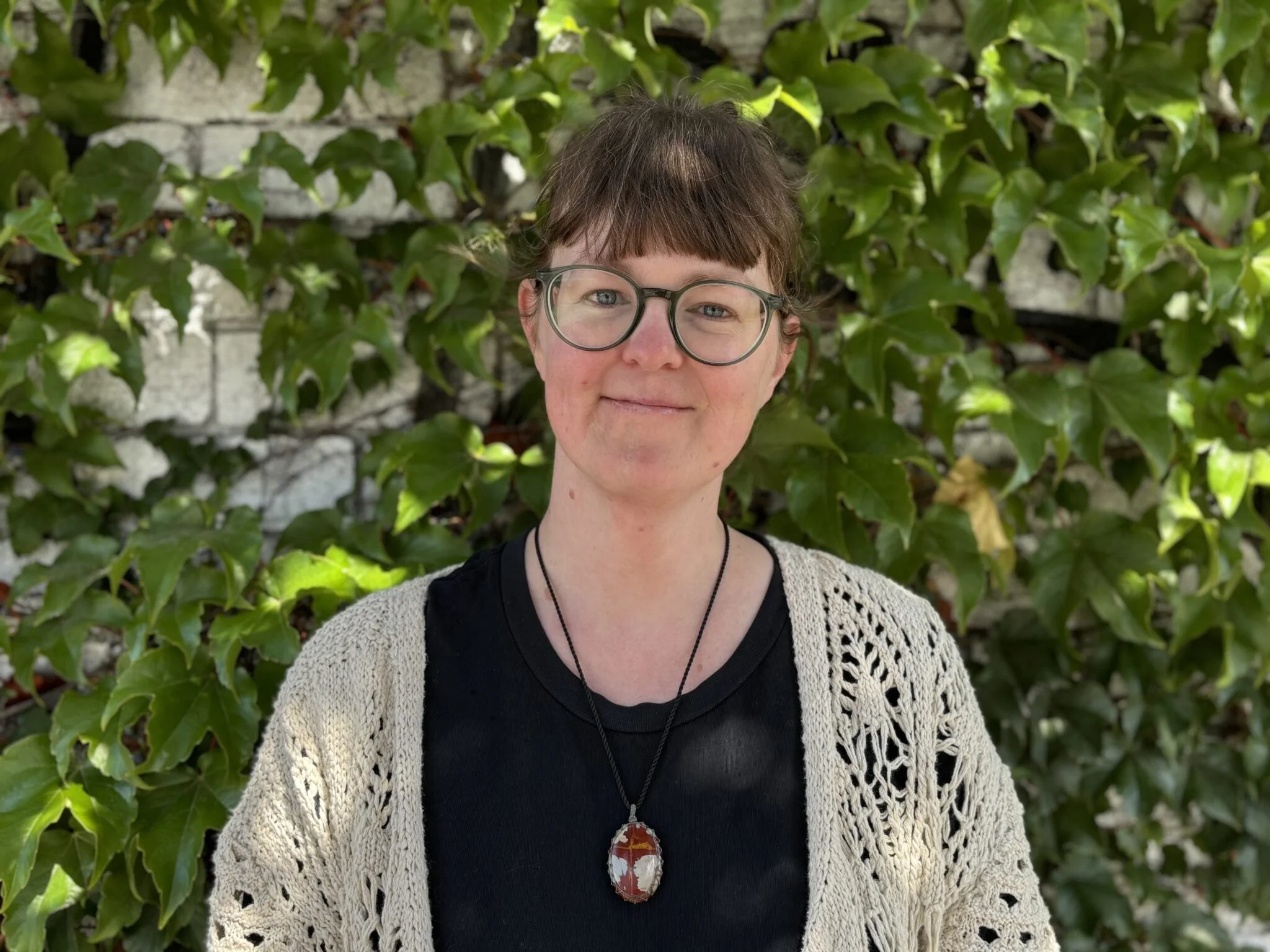 A woman with short brown hair, glasses, and light skin smiling outdoors, standing in front of green ivy and a brick wall.