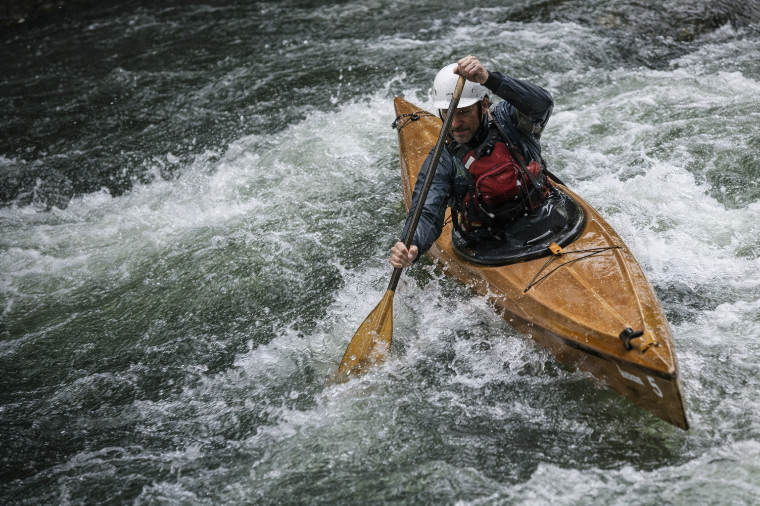 A man kayaking on turbulent river water, wearing a white helmet, a red life vest, and a black waterproof jacket.