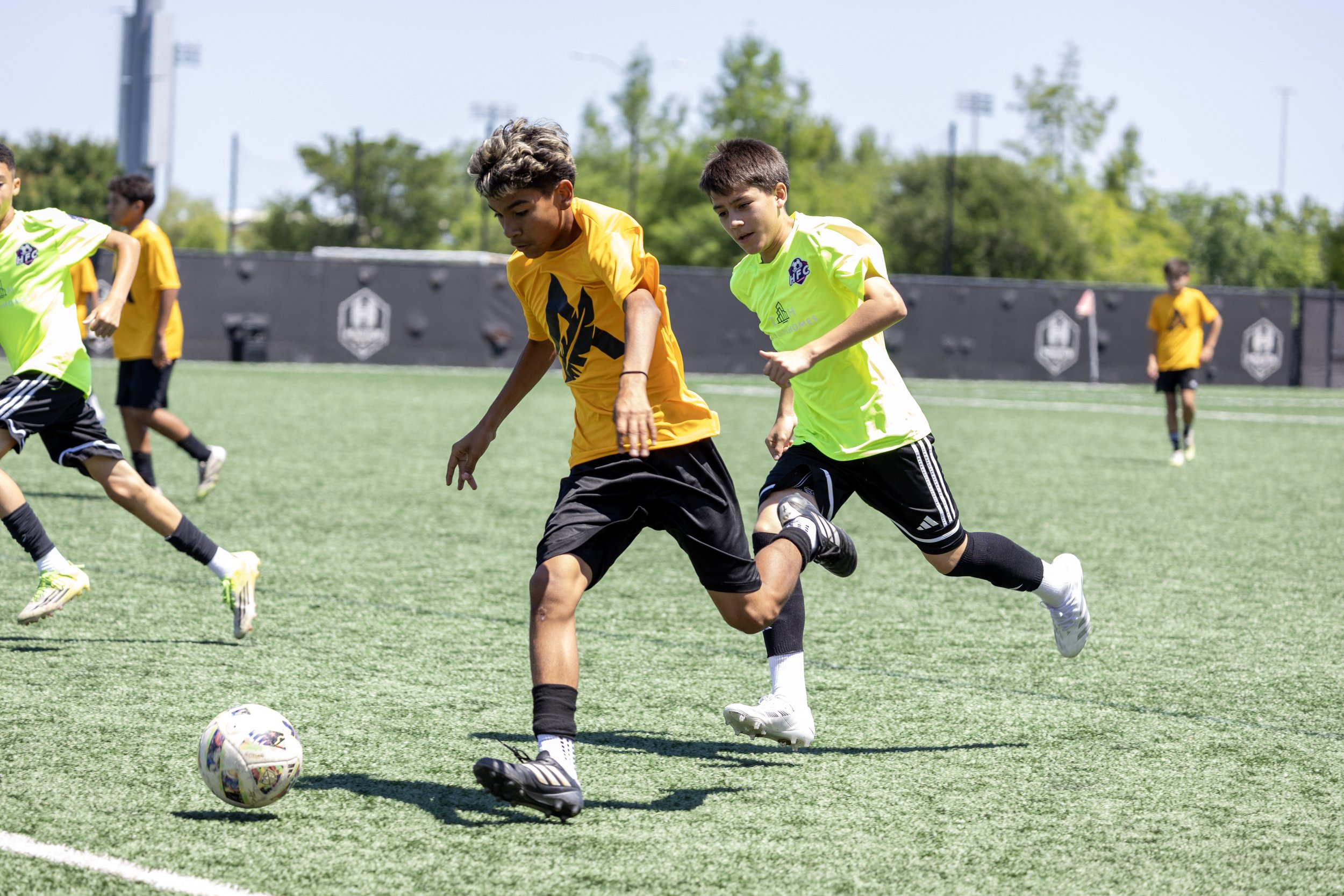 Young boys playing soccer on an outdoor field during the daytime; two boys in yellow and green jerseys chase the soccer ball.