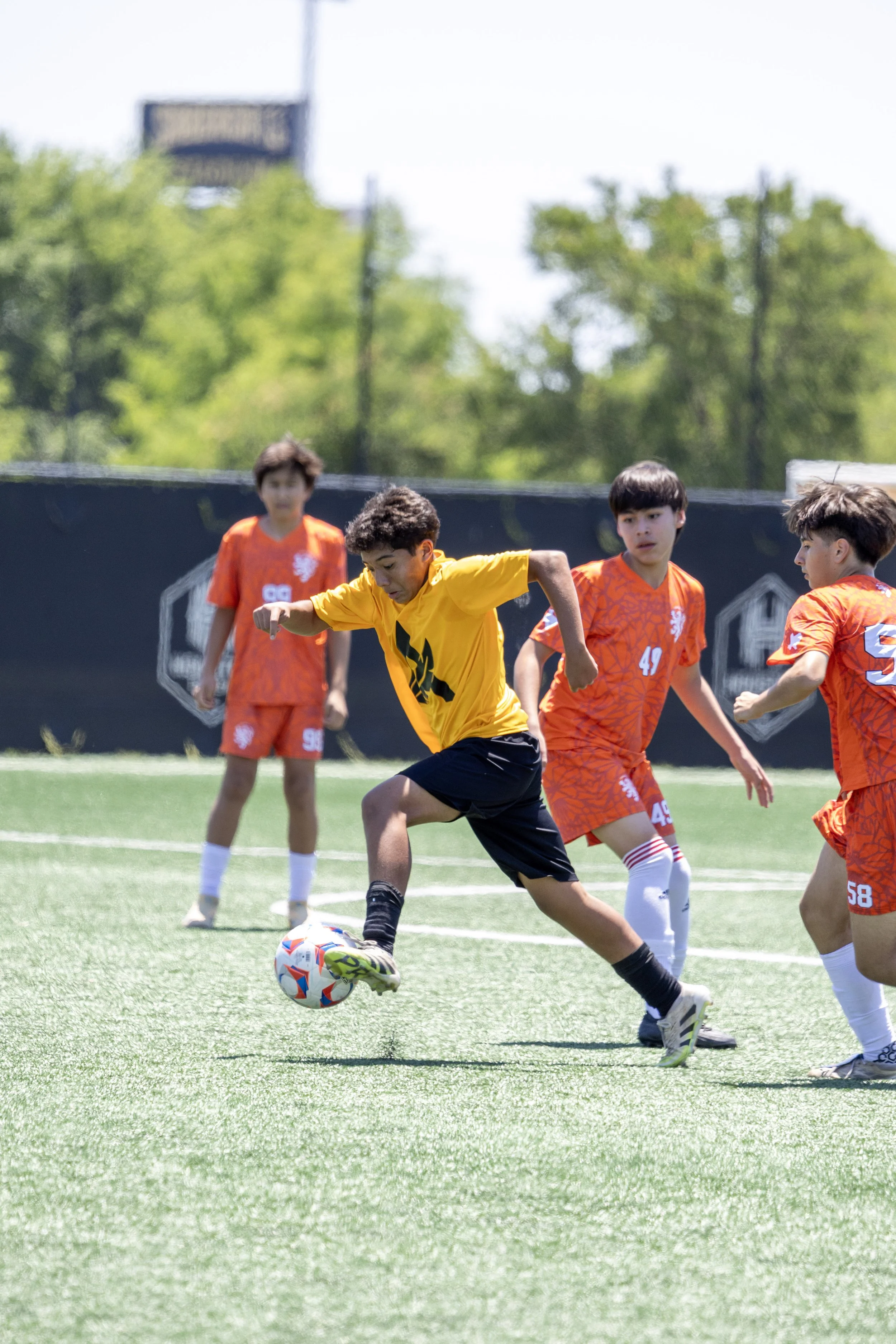 Young boys playing soccer on a field, with one boy in a yellow shirt controlling the ball as others in orange shirts watch.