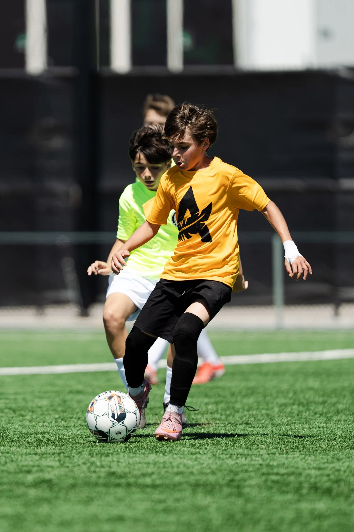 Young boys playing soccer on an outdoor field, one in a yellow jersey and black shorts controlling the ball, while others chase.