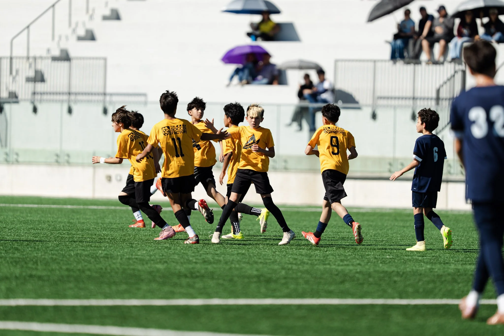 Kids playing soccer on a bright green field with spectators watching from stands, some holding umbrellas.