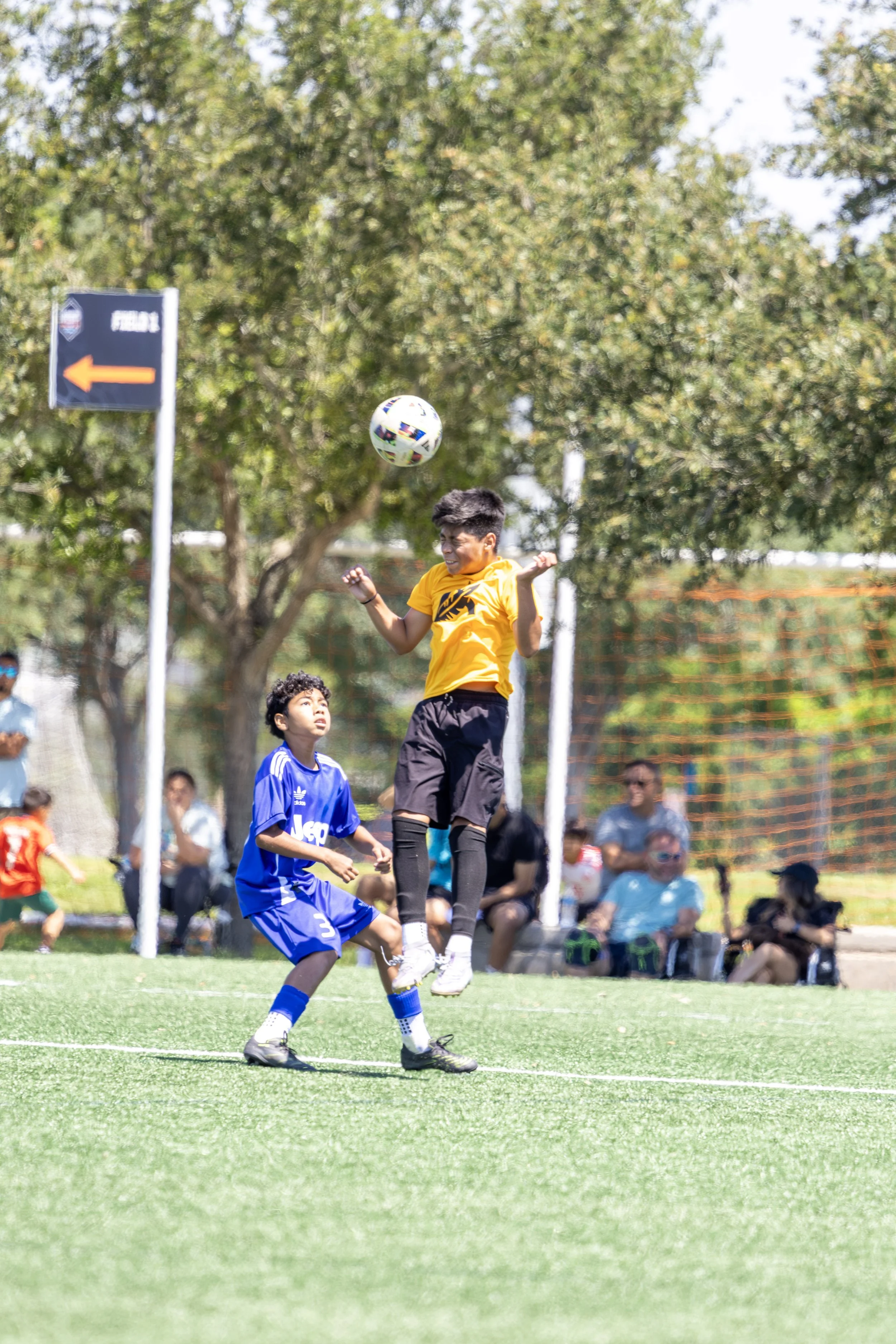 Two boys playing soccer on a field, with spectators watching in the background. One boy in a yellow shirt jumps to head the ball, while the other boy in a blue jersey stands nearby.