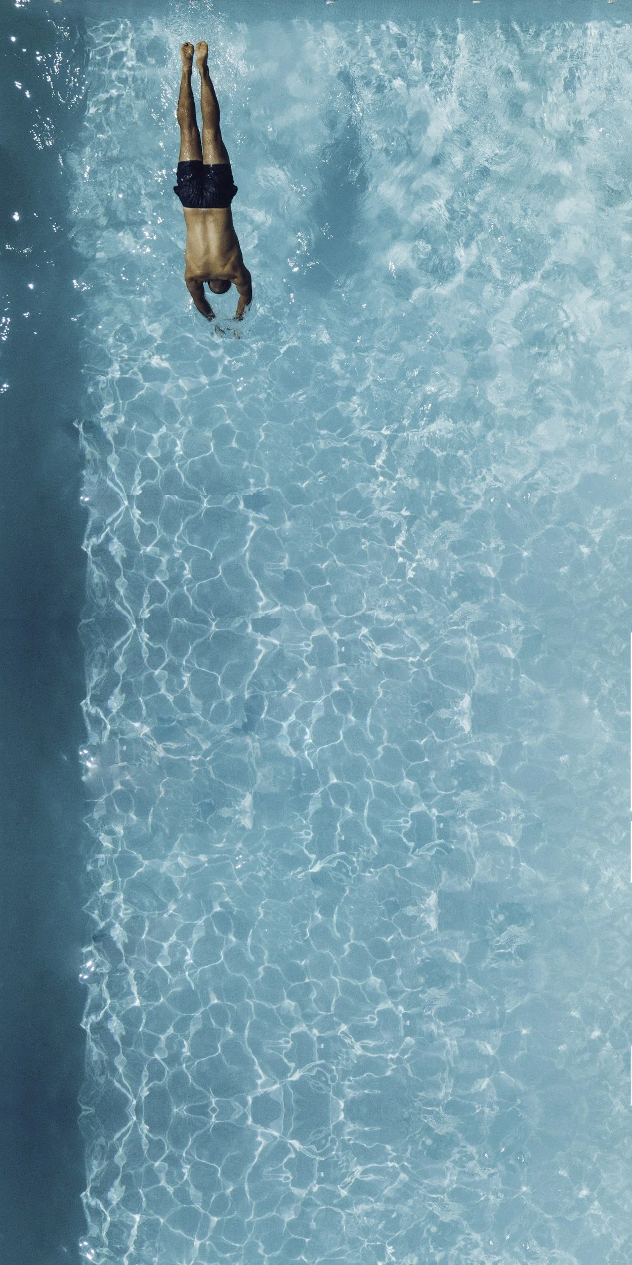 A person diving into a pool with clear blue water, wearing black swimming trunks.