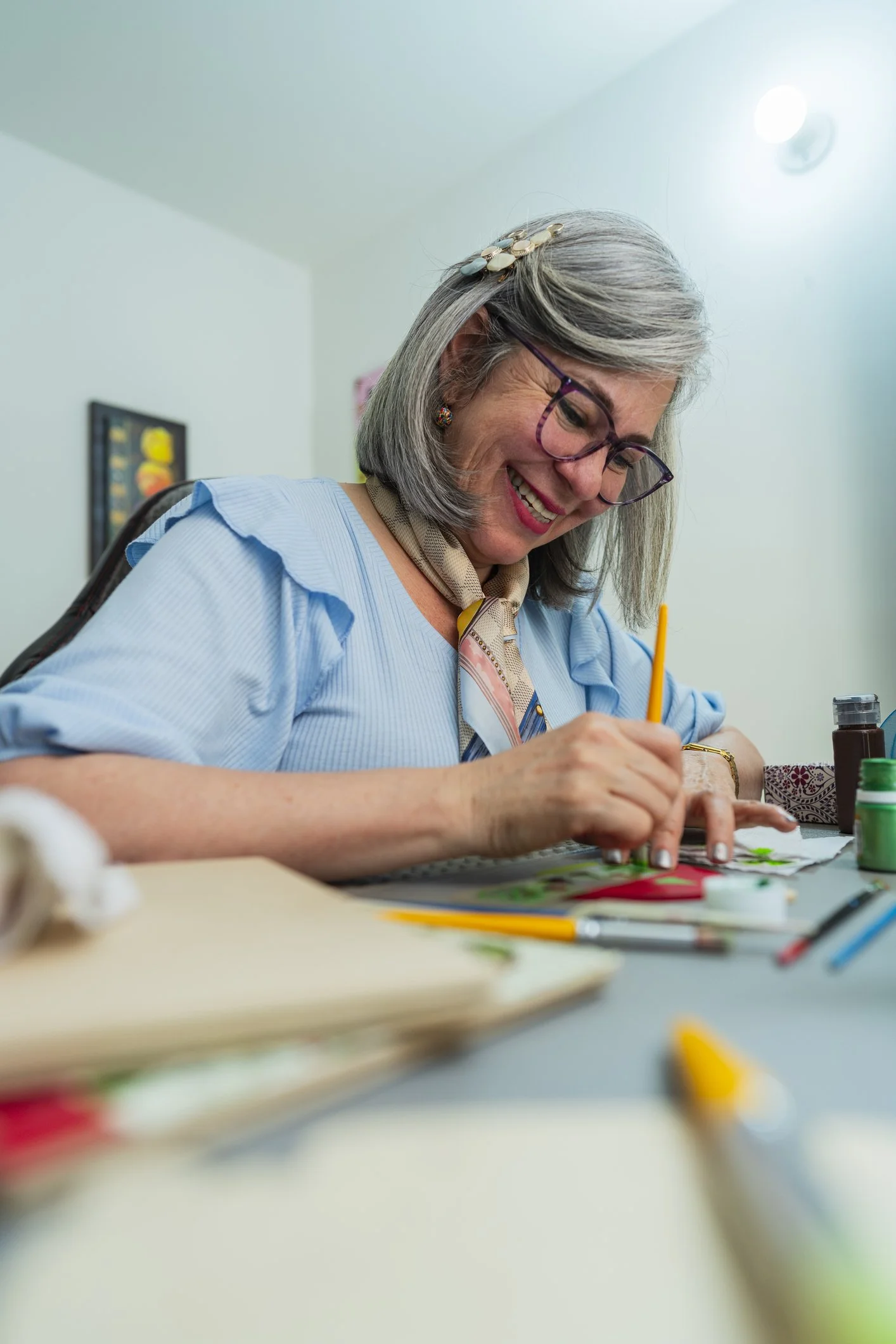 A woman with gray hair, glasses, and colorful earrings, smiling while painting or drawing at a desk with art supplies.