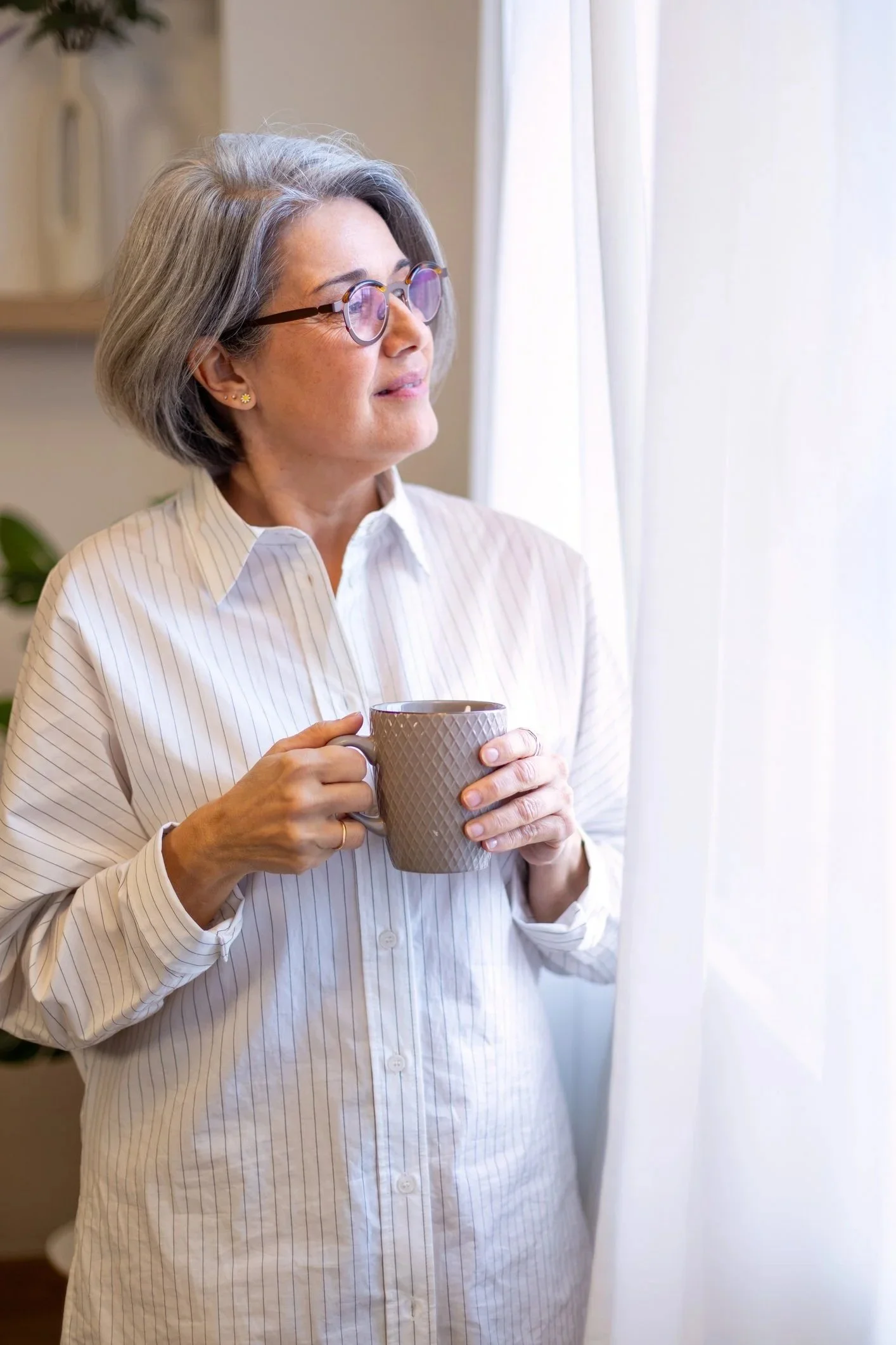 A woman with gray hair, glasses, and earrings holding a beige textured mug near a window with white curtains.