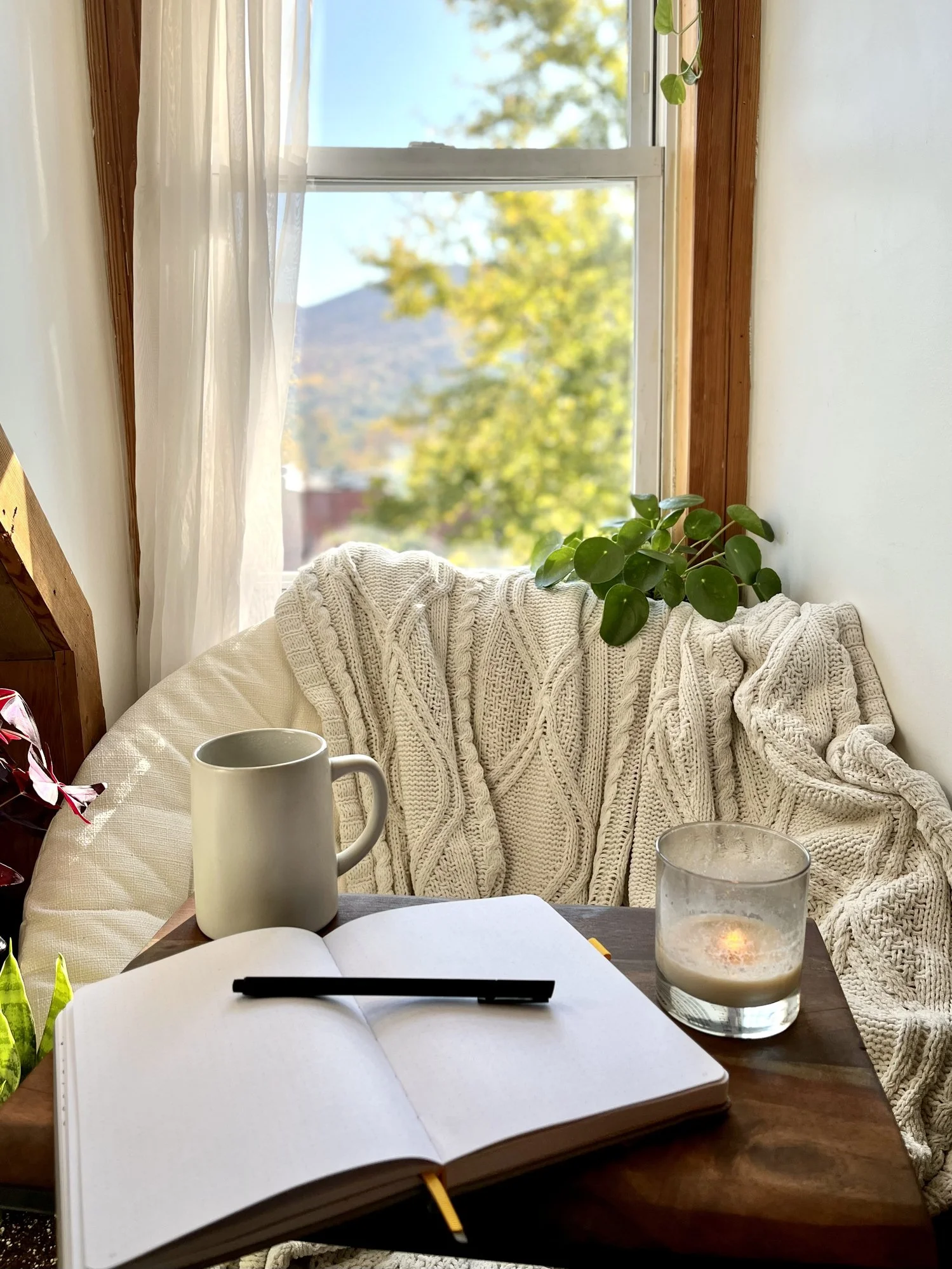 Cozy window nook with blanket, candle, notebook, and coffee mug in warm natural light.