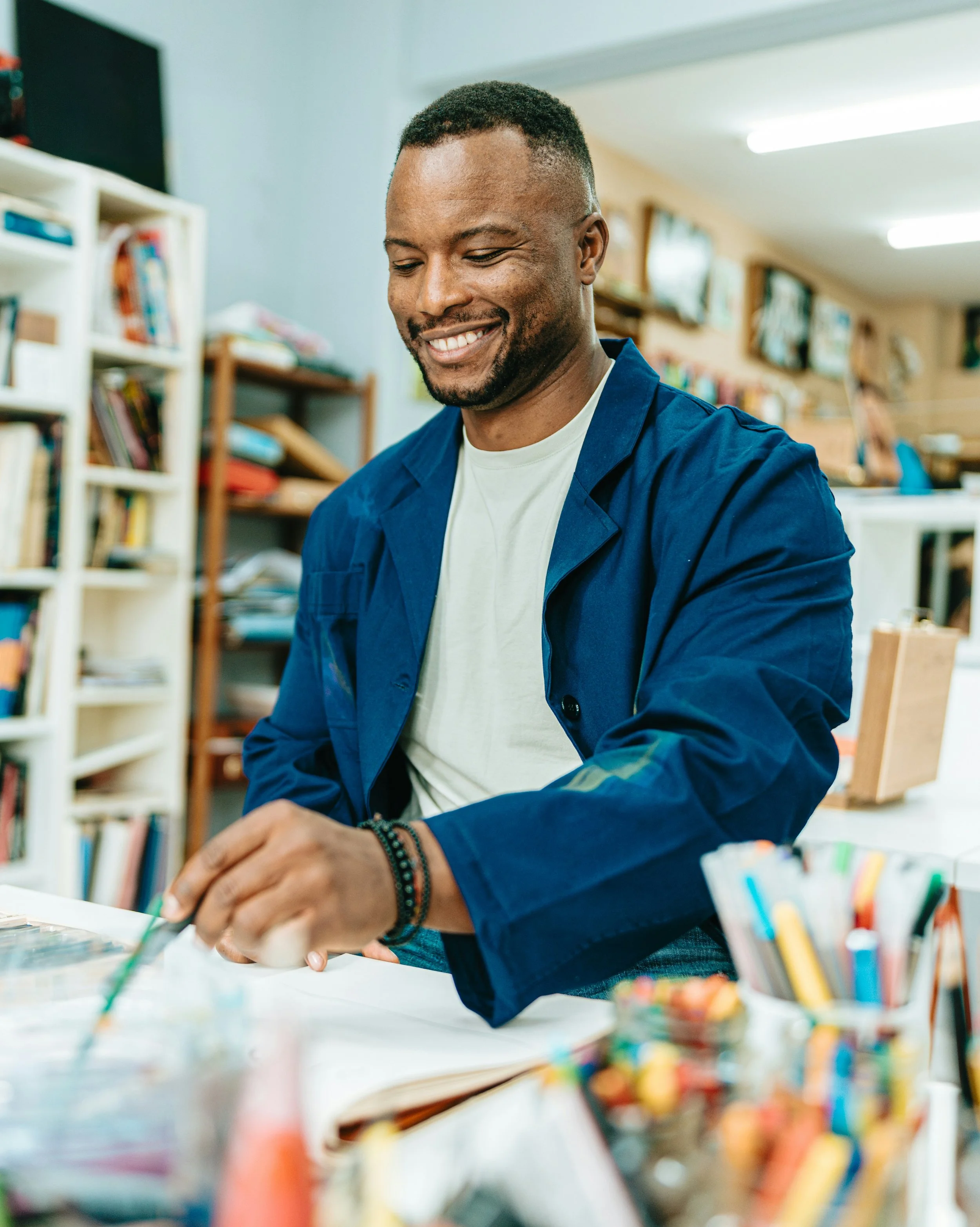 A man smiling while working in a craft or art studio surrounded by colorful art supplies and books.