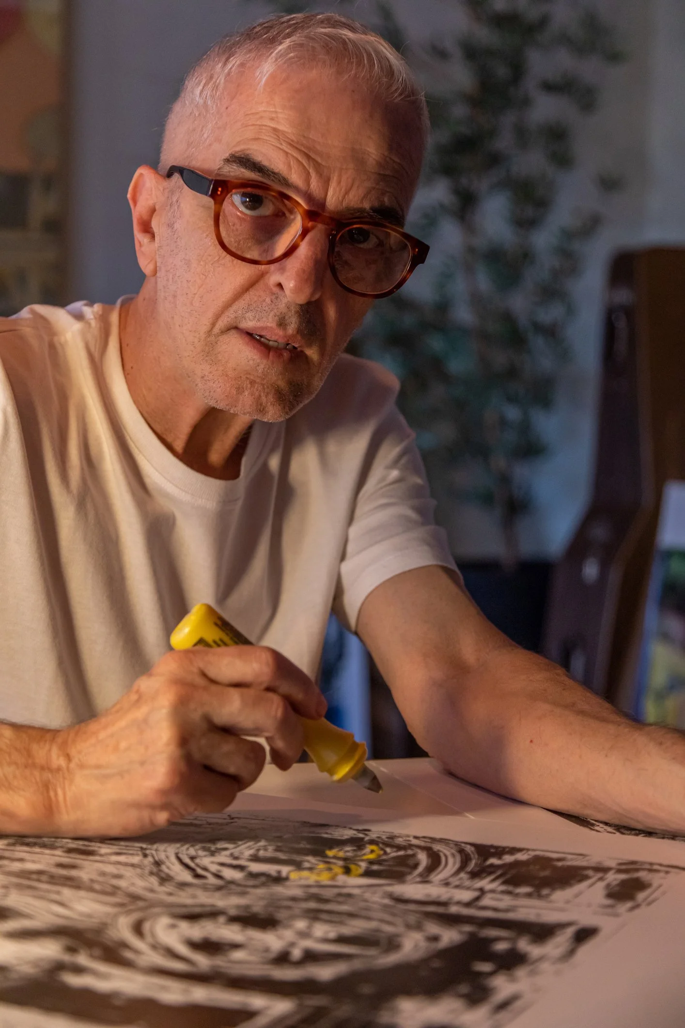 An older man with glasses and a white t-shirt sitting at a table, using a yellow tool to carve or etch a detailed black and white artwork on a large sheet of paper.