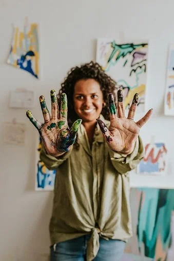 A woman smiling with hands covered in colorful paint, standing in front of abstract paintings on a wall.