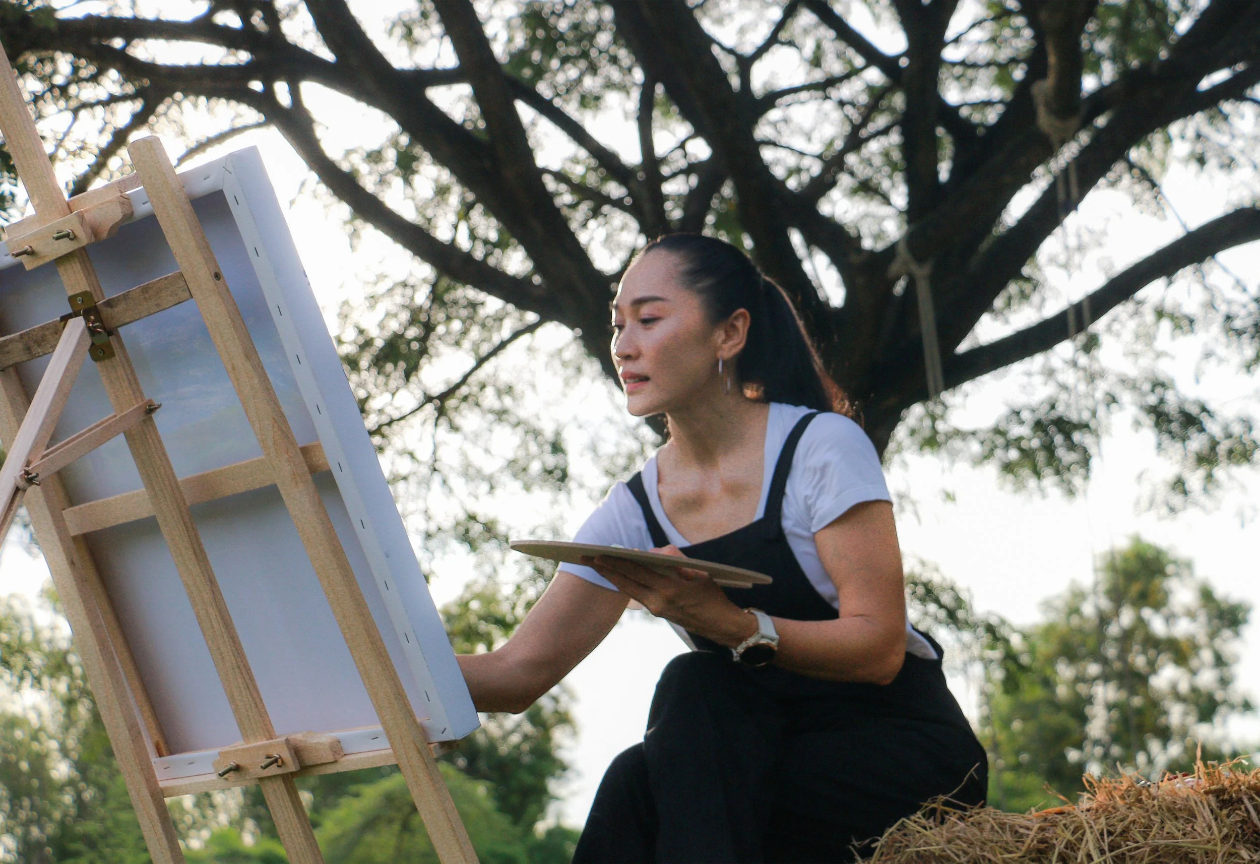 A woman painting outdoors on a canvas set on an easel, with trees in the background.