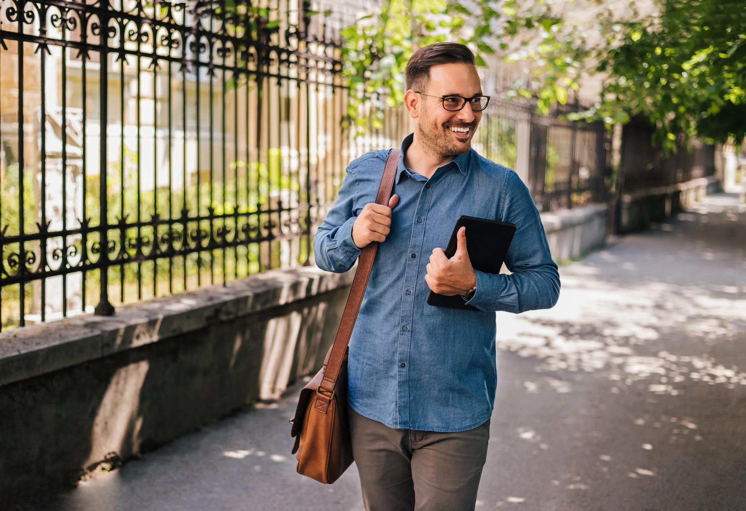 A smiling man with glasses wearing a blue button-up shirt and carrying a brown shoulder bag, walking outdoors on a sidewalk next to a decorative black metal fence and green trees.