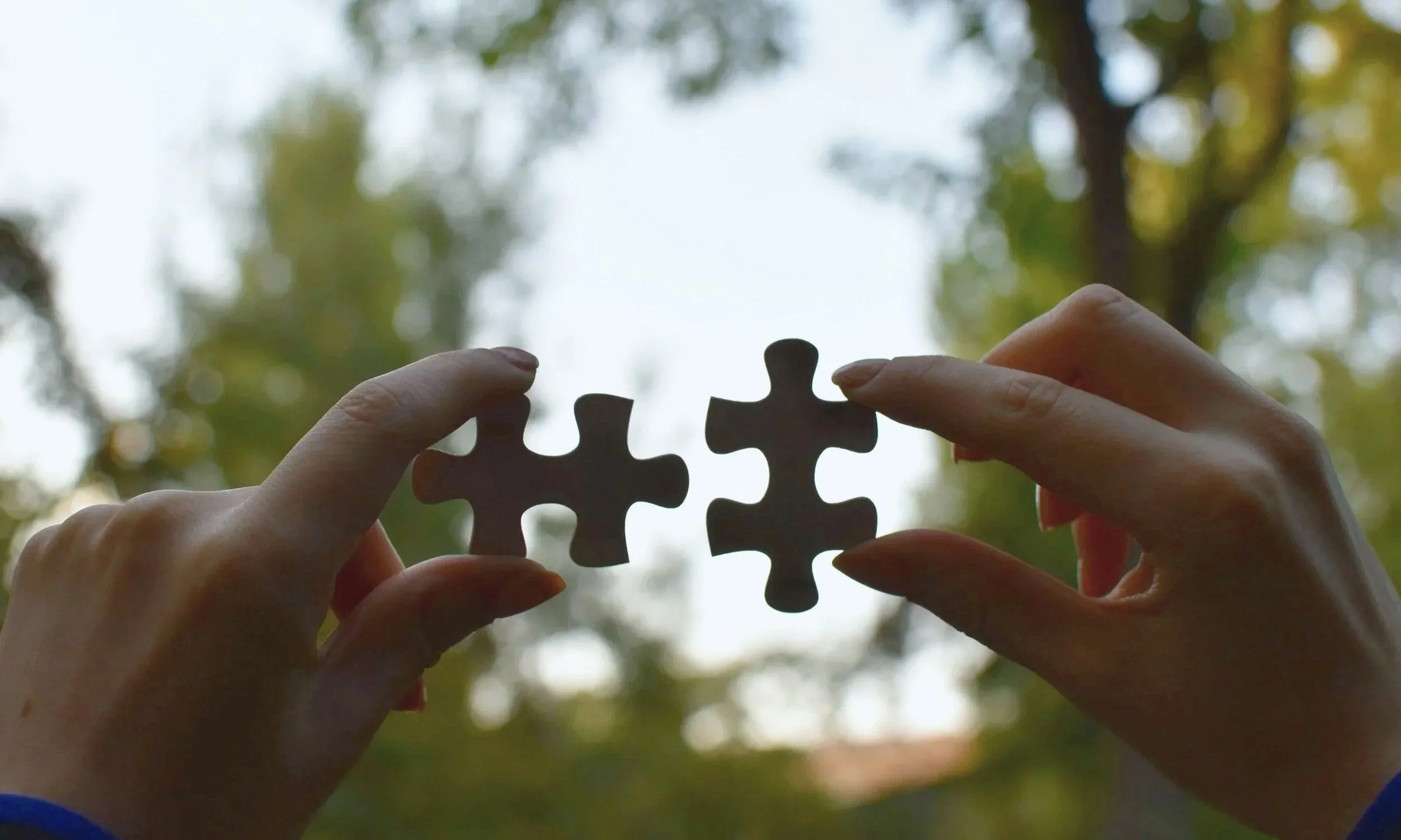 Hands holding two puzzle pieces against a soft outdoor background, symbolizing connection and problem-solving.