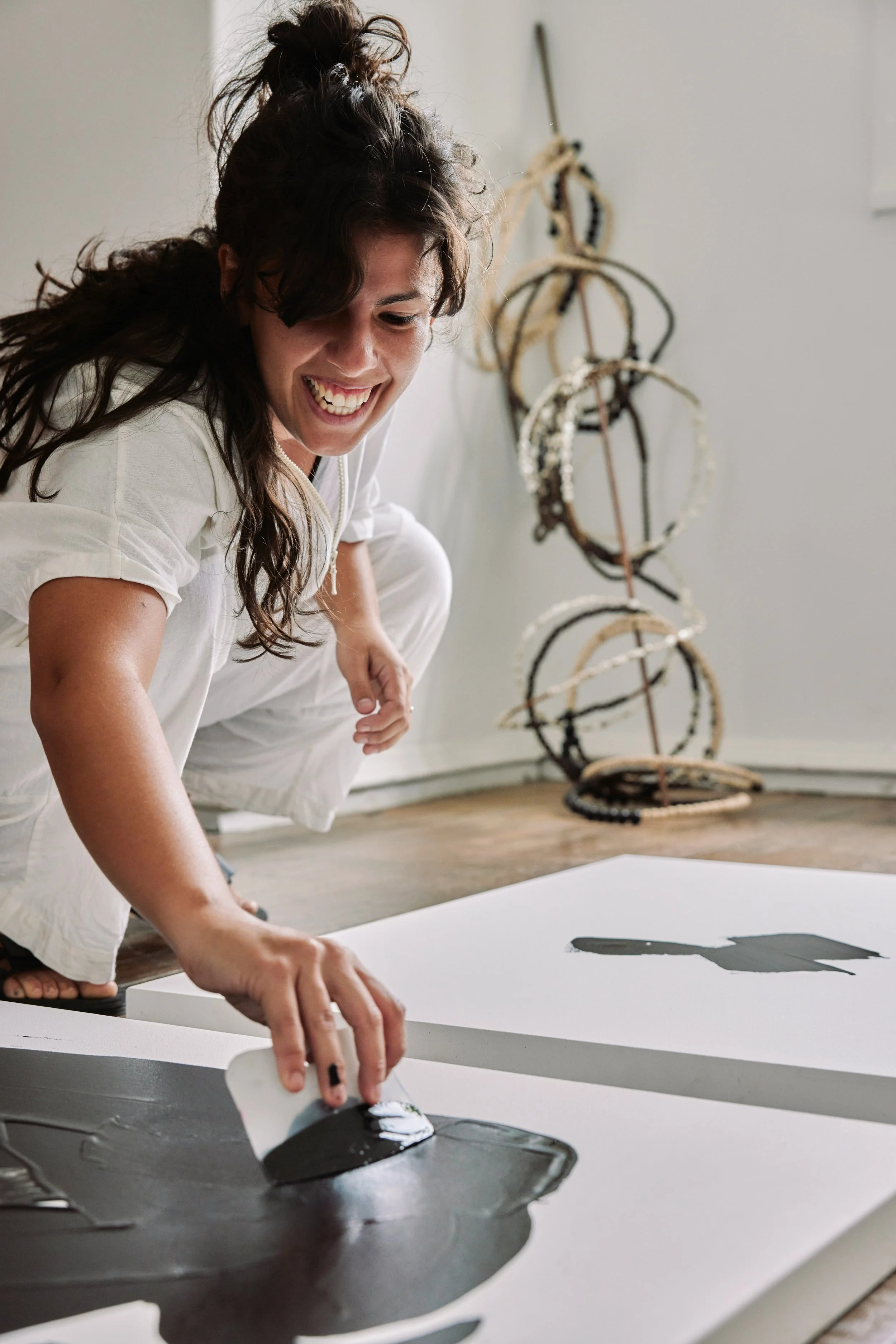 A woman smiling and working on a large white canvas, spreading black paint with a spatula. In the background, there are artistic necklaces hanging on a wall.