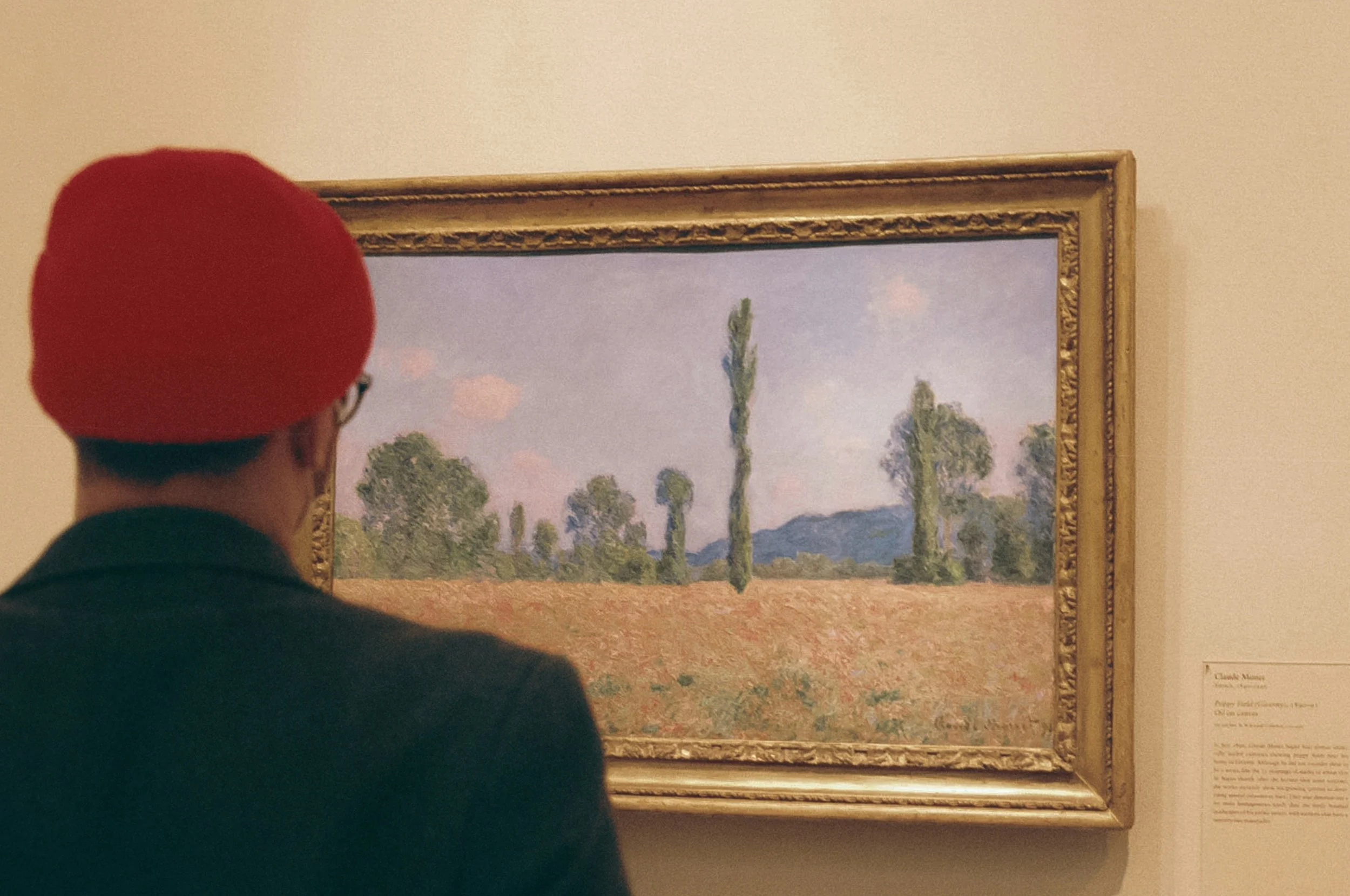 Person wearing a red beret and black jacket viewing a landscape painting of trees and a field.