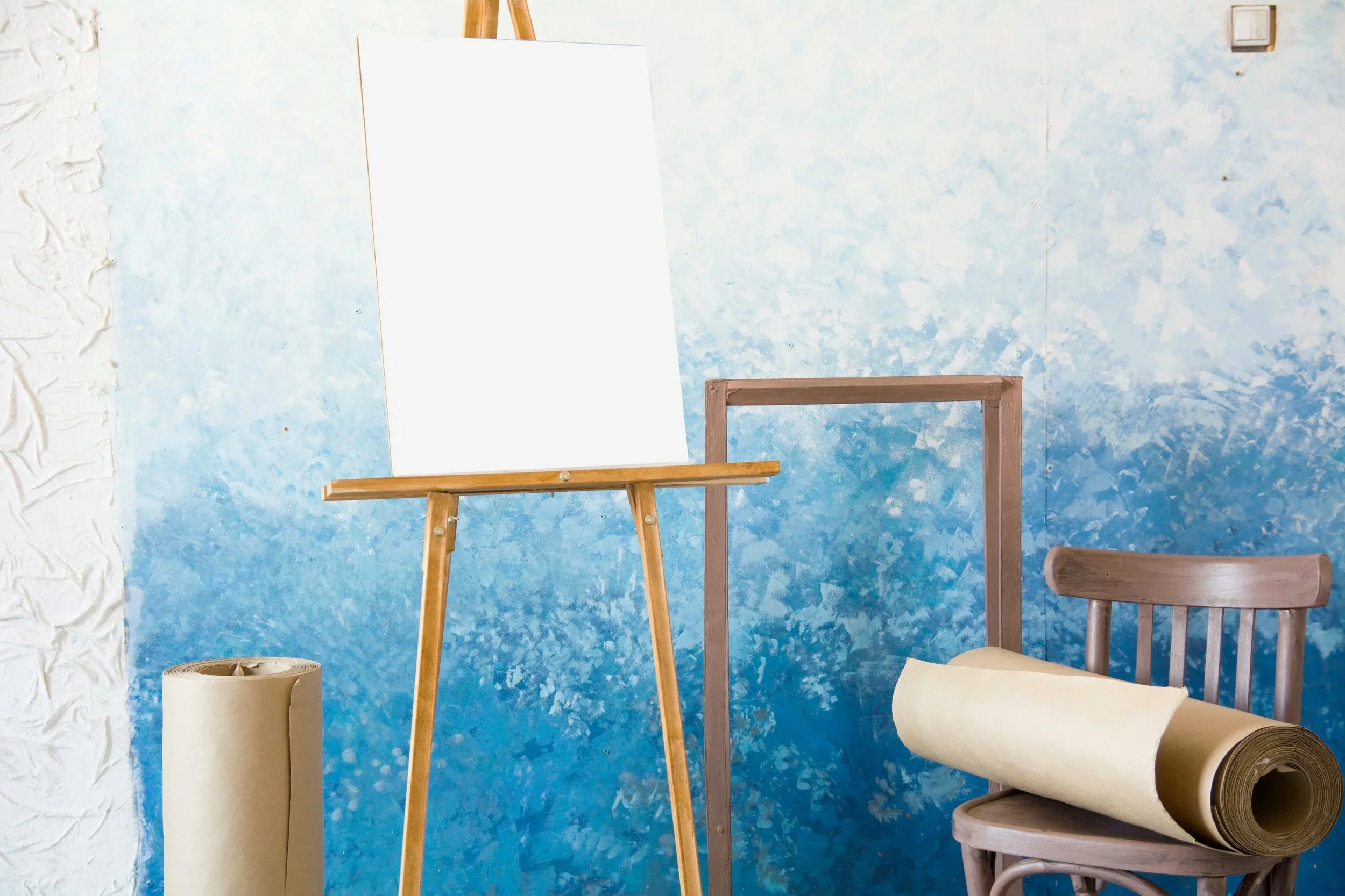 An art studio corner with an easel holding a blank white canvas, a wooden chair with a roll of wallpaper or canvas on it, a small wooden frame or stand, and a roll of beige paper or canvas on the floor, against a blue, textured wall with a white section on the left.