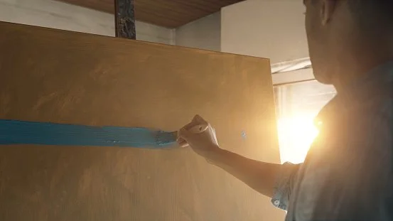 Person painting a horizontal blue stripe on a brown wall.