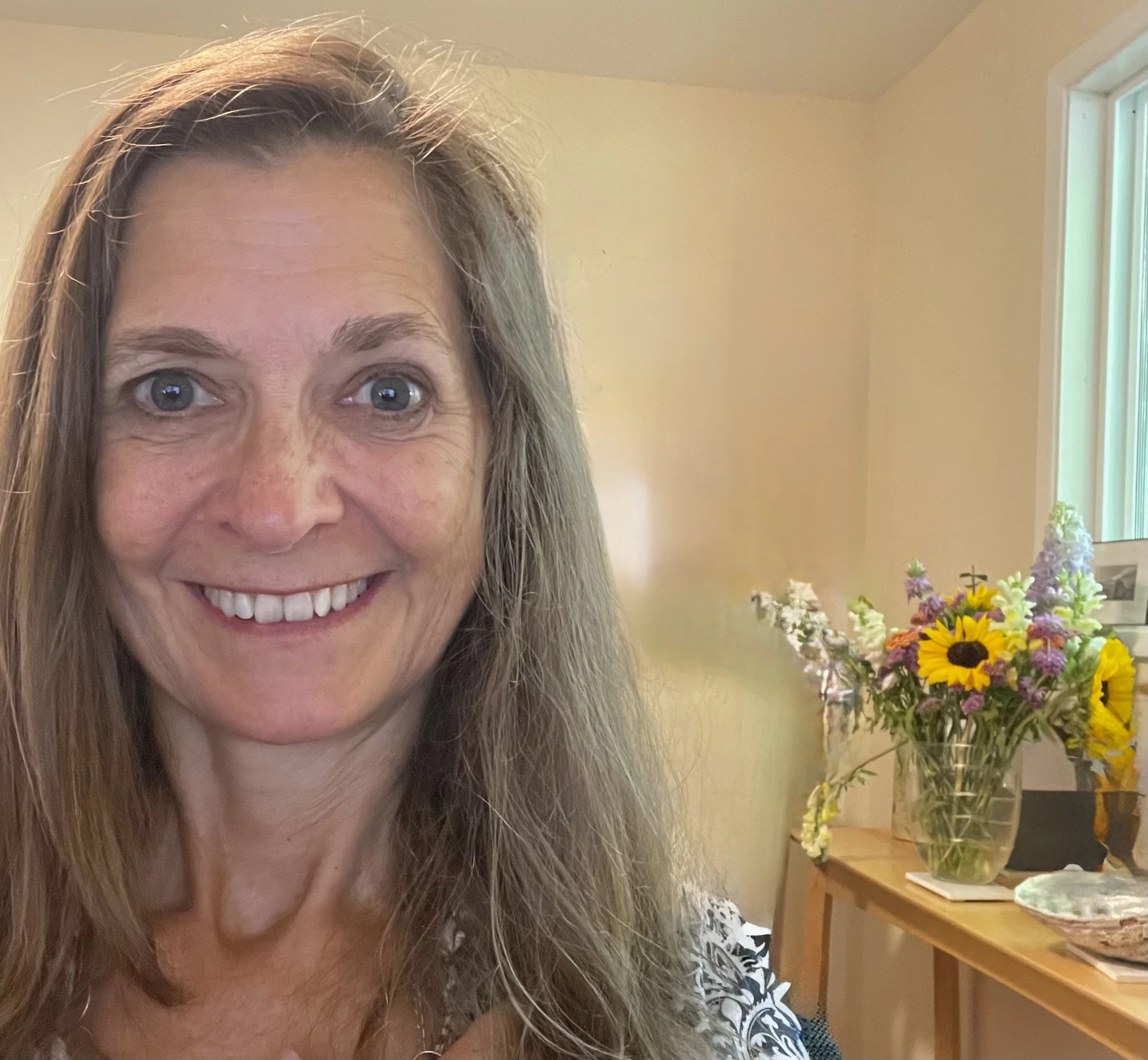 A woman with long, wavy, light brown hair smiling at the camera in a room with a table holding a large vase of colorful flowers, including sunflowers and purple and white blooms. A window with vertical blinds is in the background.